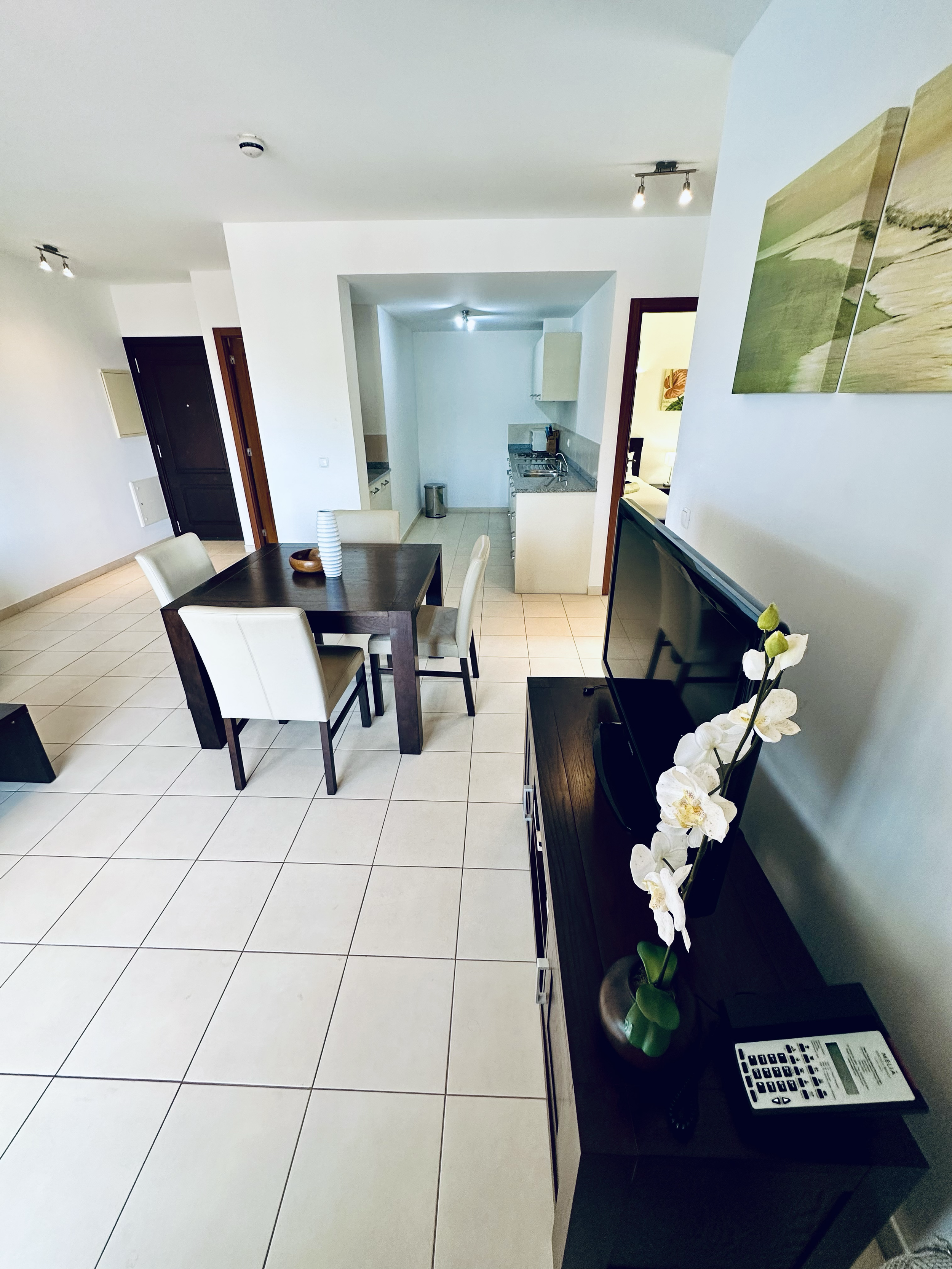 Interior of a modern apartment or hotel room with a small dining area, kitchen, and a hallway. The room features white tiled flooring, a dark wood table with cream-colored chairs, wall art, a sideboard with a vase of white orchids, a TV, and some ele