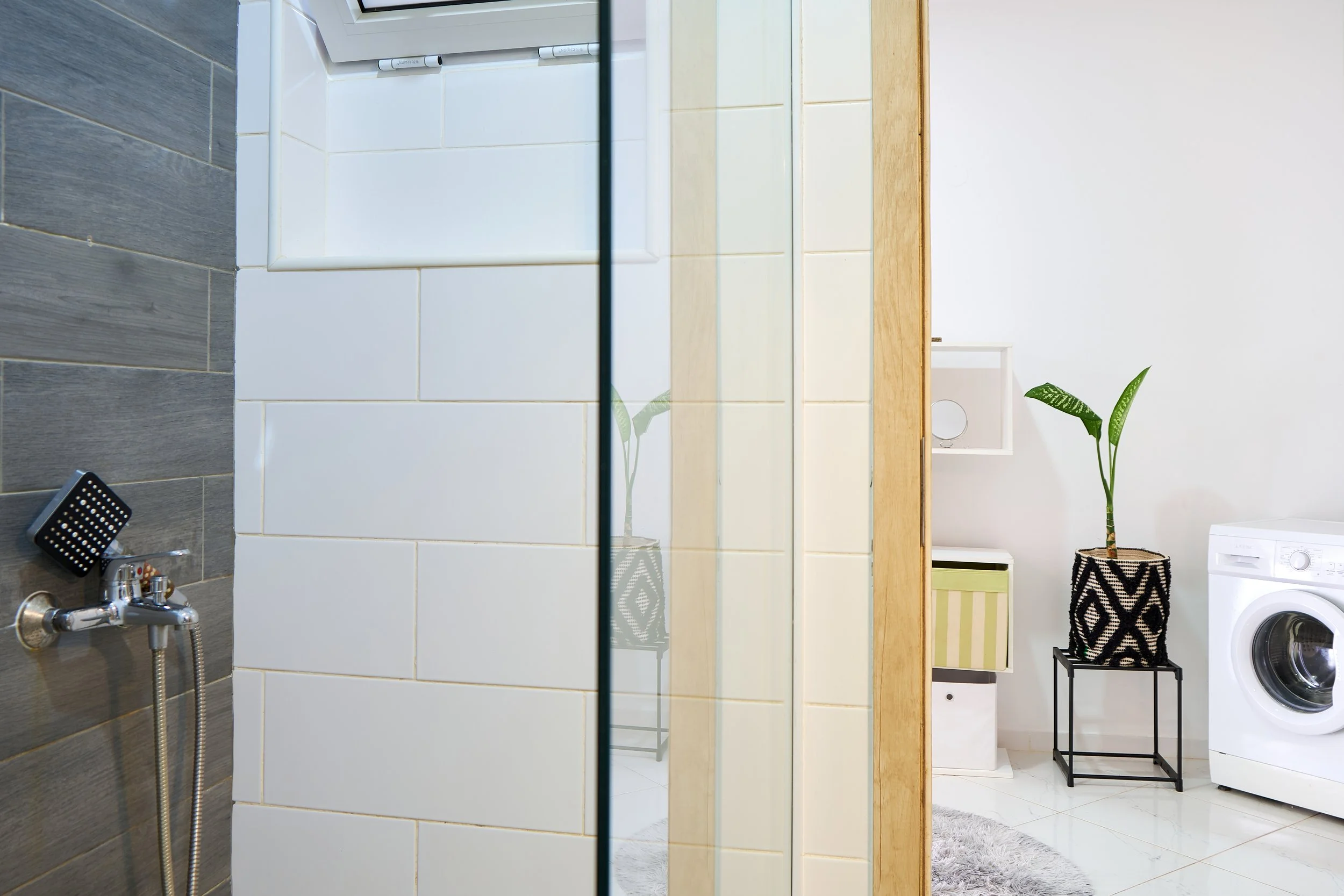 Modern bathroom with a shower and a view into an adjacent laundry area featuring a washing machine, potted plant, storage baskets, and a white wall.