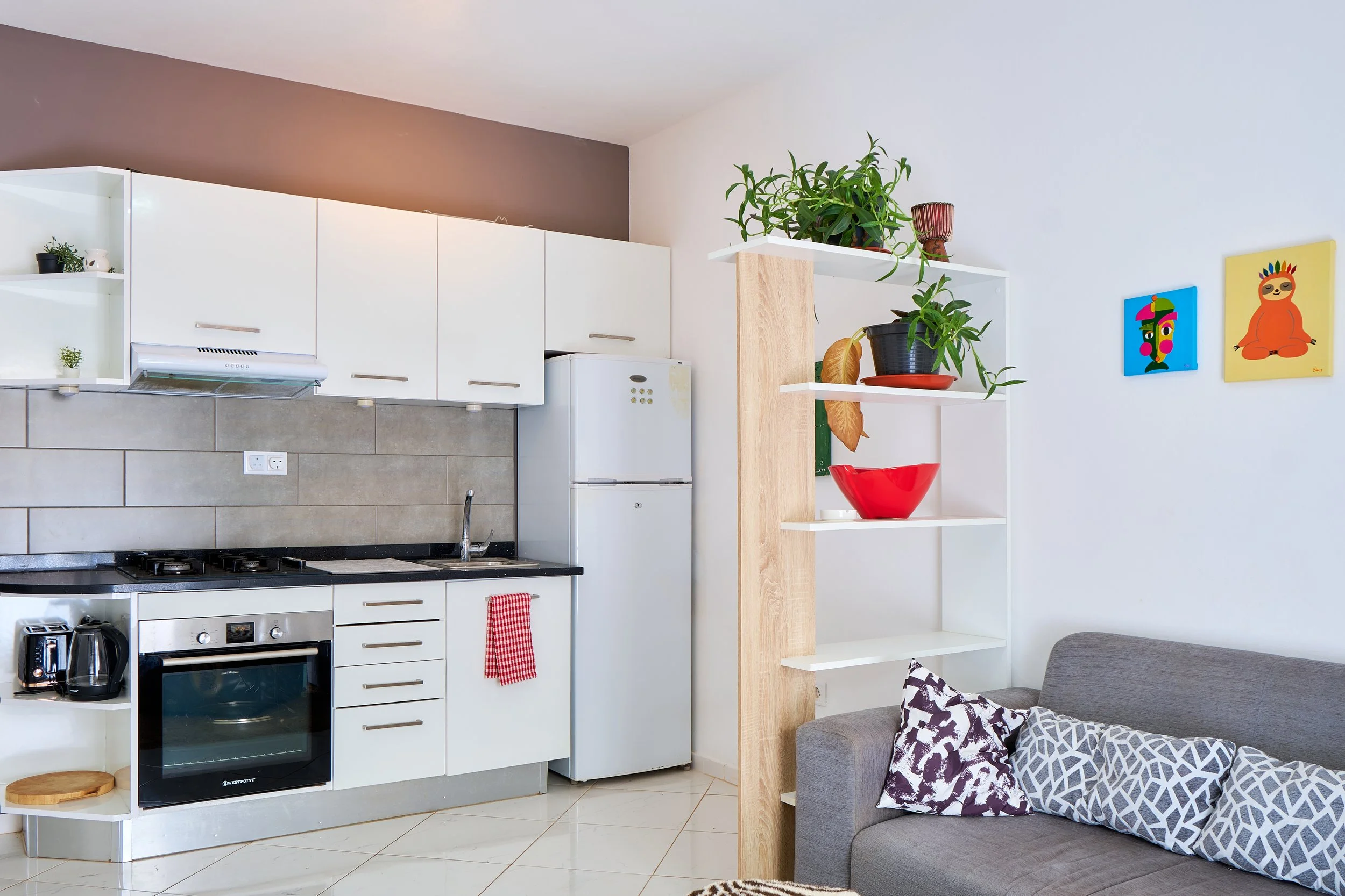 Interior view of a kitchen with white cabinets, a stove, refrigerator, and small potted plants on open shelves. Part of a living room with a gray sofa and decorative pillows is visible.