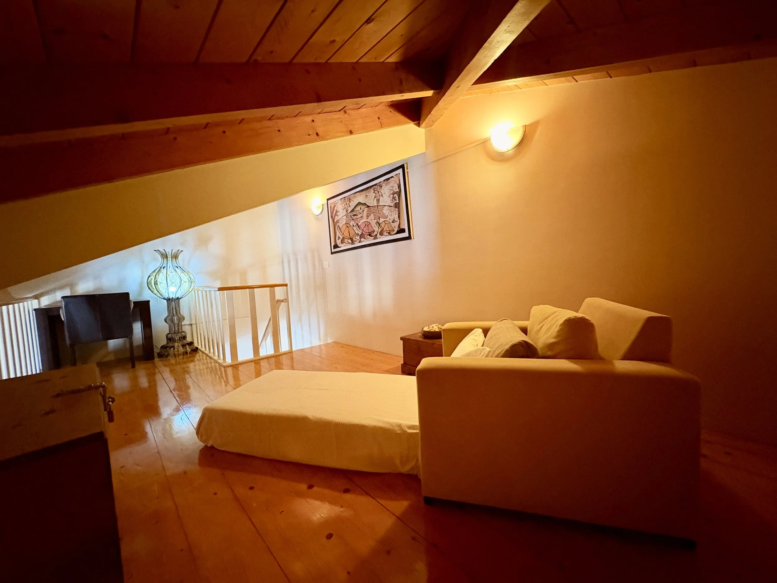 Interior of a room with wooden beams on the ceiling, a yellow wall with a framed picture, a table lamp, and beige sofa, illuminated by wall lights.