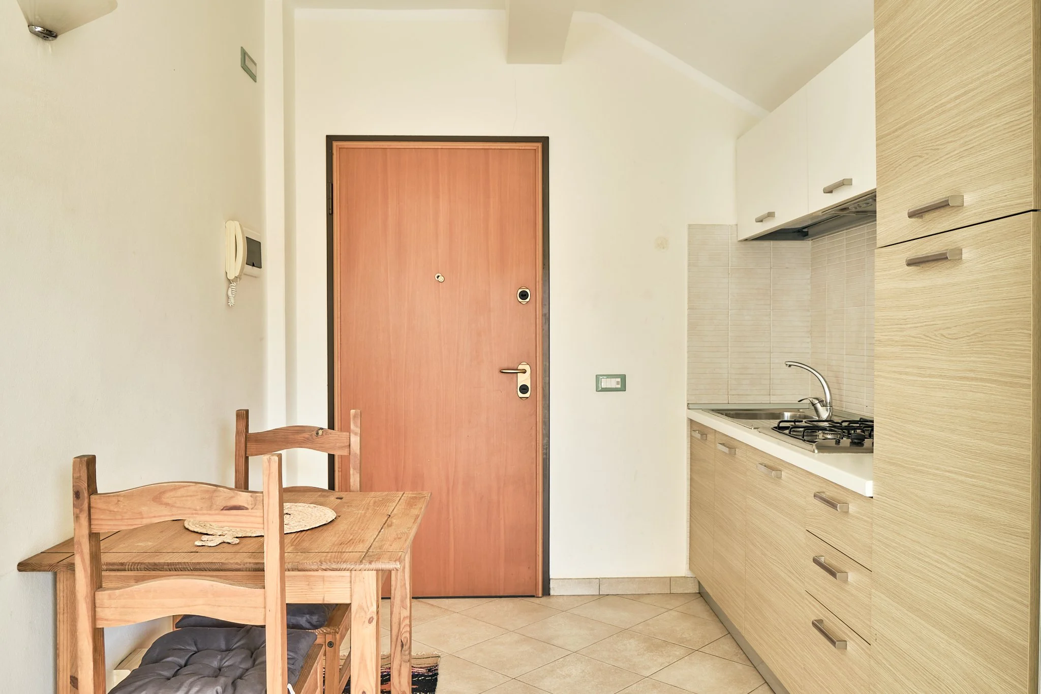 A small kitchen area with a wooden dining table and chairs, a pink front door, and white cabinetry with a stovetop and sink. An intercom is on the wall next to the door.