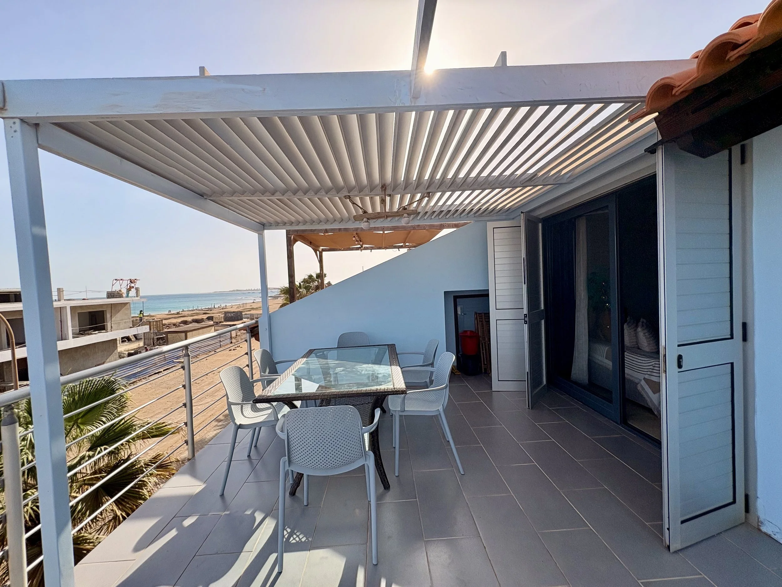 Balcony with a glass-top table and white chairs overlooking a beach and ocean, with a white pergola and a sliding glass door.