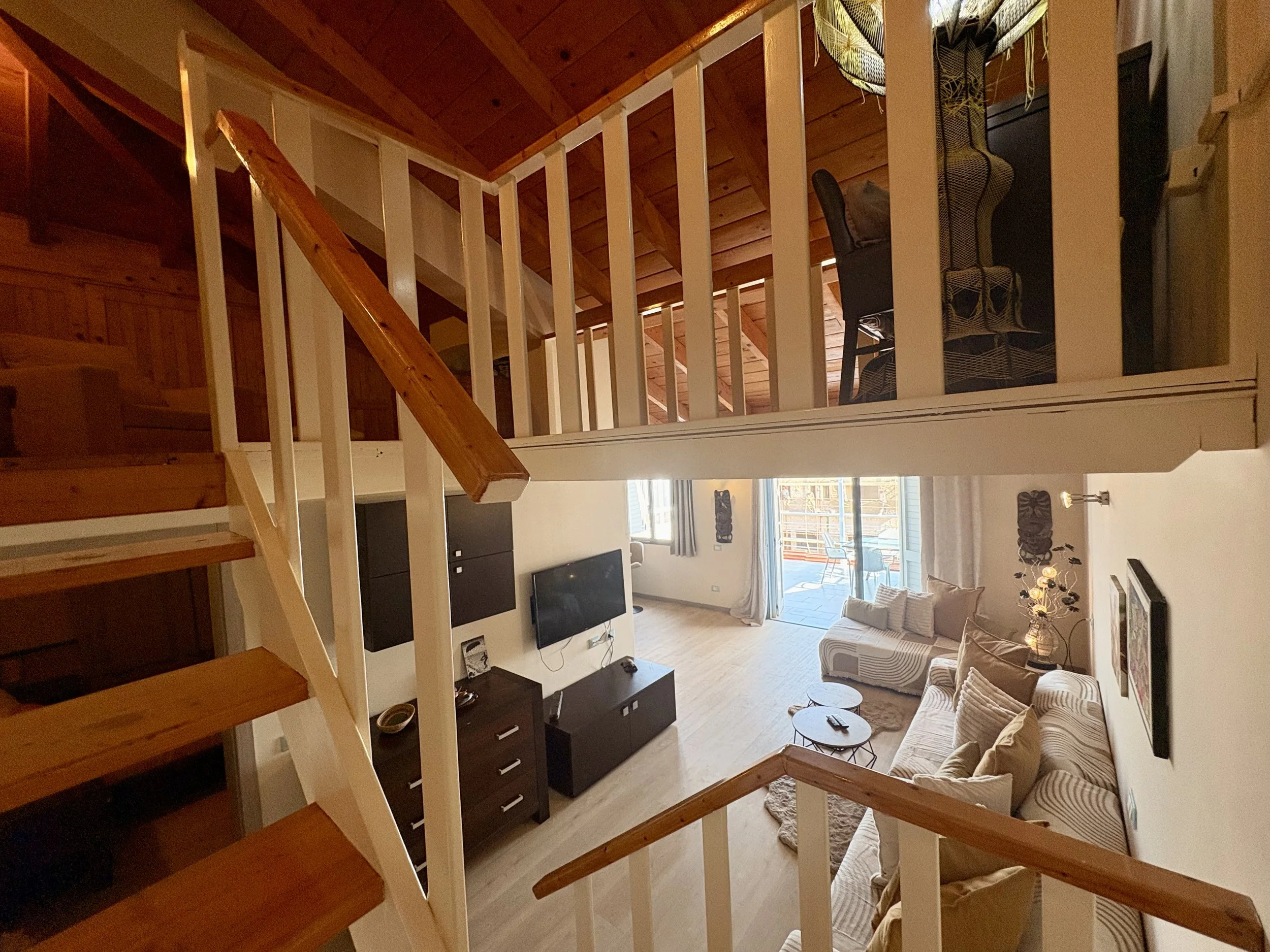 Interior view of a living room with a staircase in the foreground, featuring wooden steps and white railing. The room has large windows, a TV mounted on the wall, and seating furniture including a patterned armchair and a striped sofa. There are deco