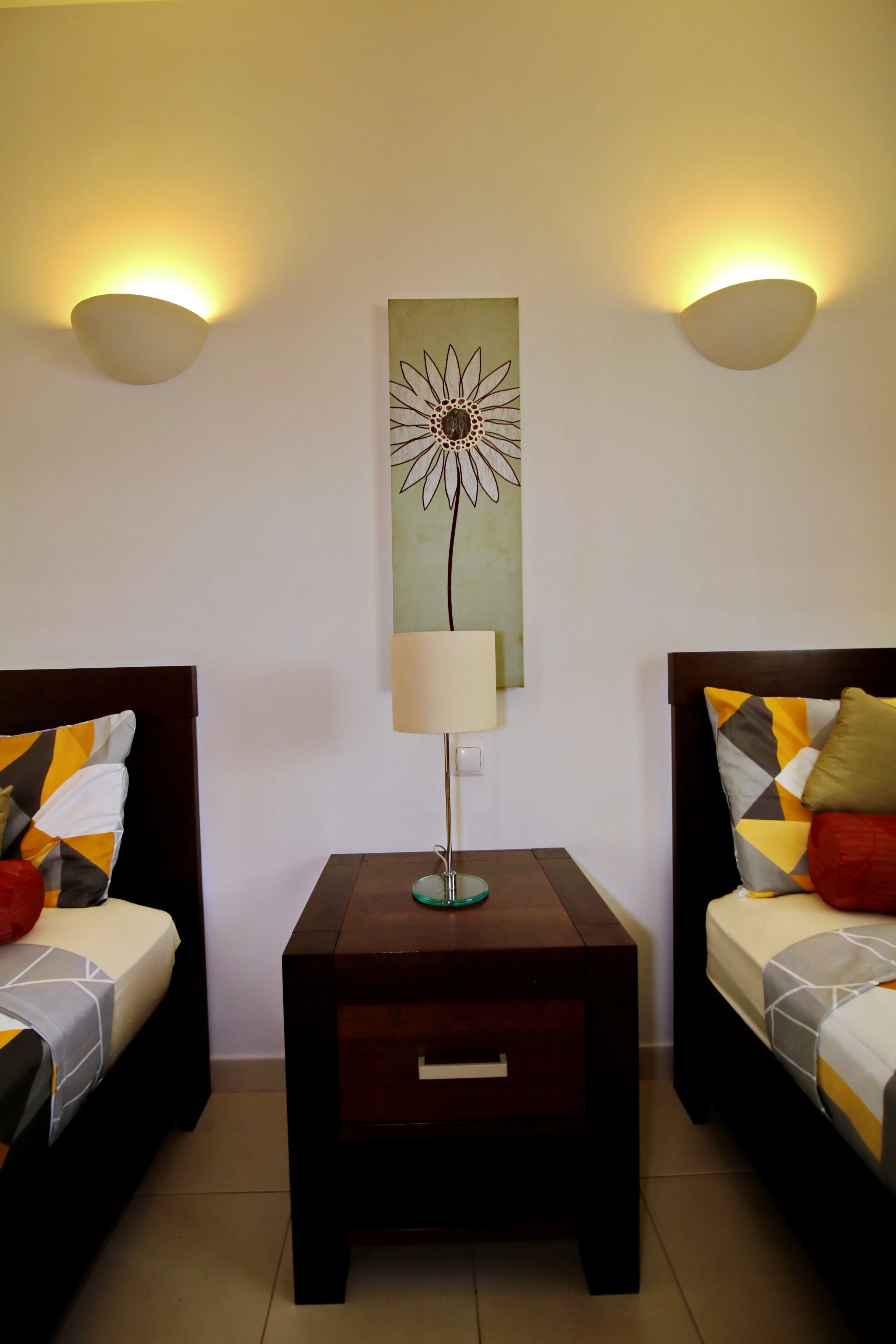 A living room corner with two single beds, a wooden nightstand with a lamp, a wall-mounted floral artwork, and wall-mounted lights illuminating the ceiling.