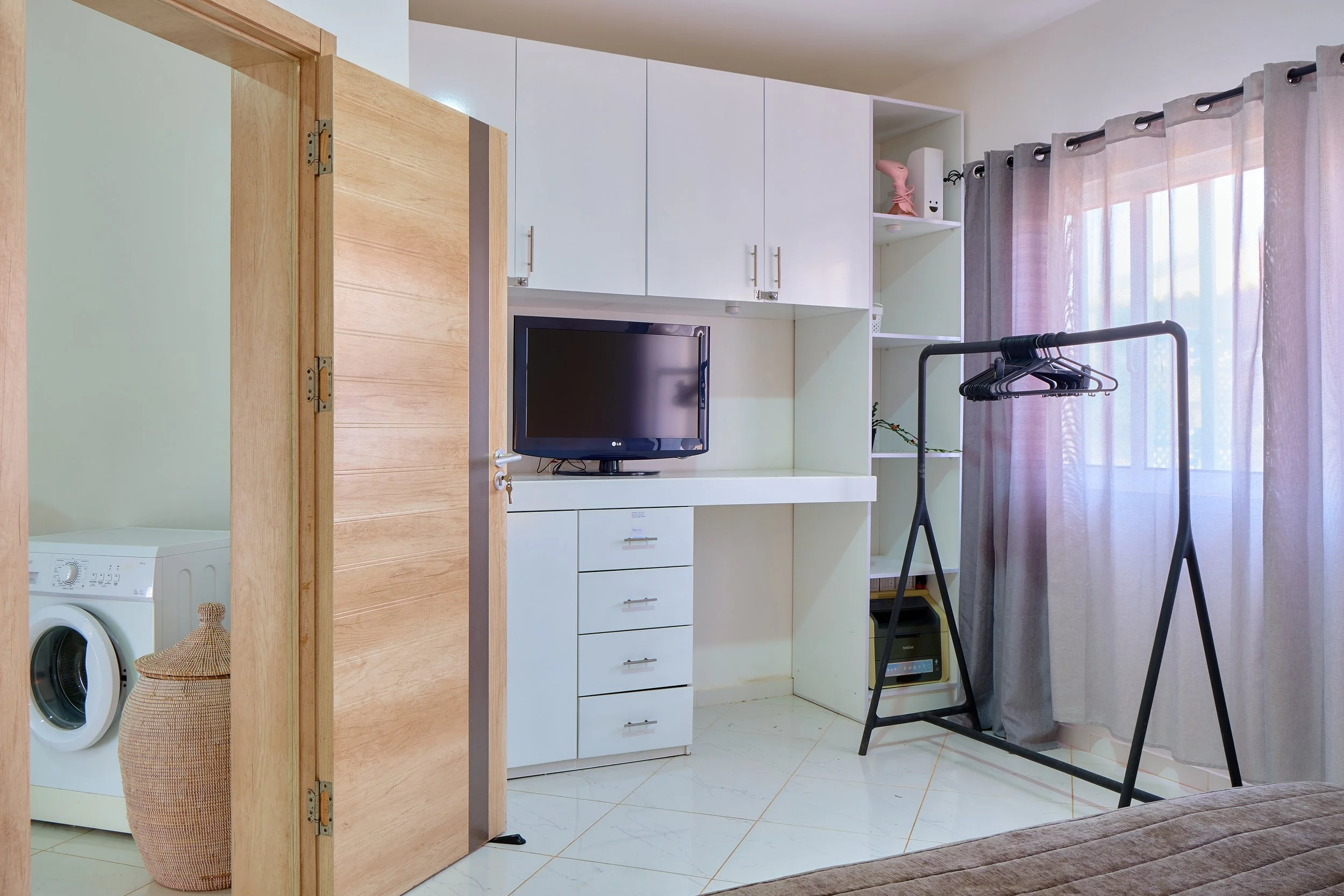 A cozy room with white shelving and cabinets, a small television, a clothes rack with hangers, a window with gray curtains, and a washing machine partially visible in a laundry area behind a wooden door.