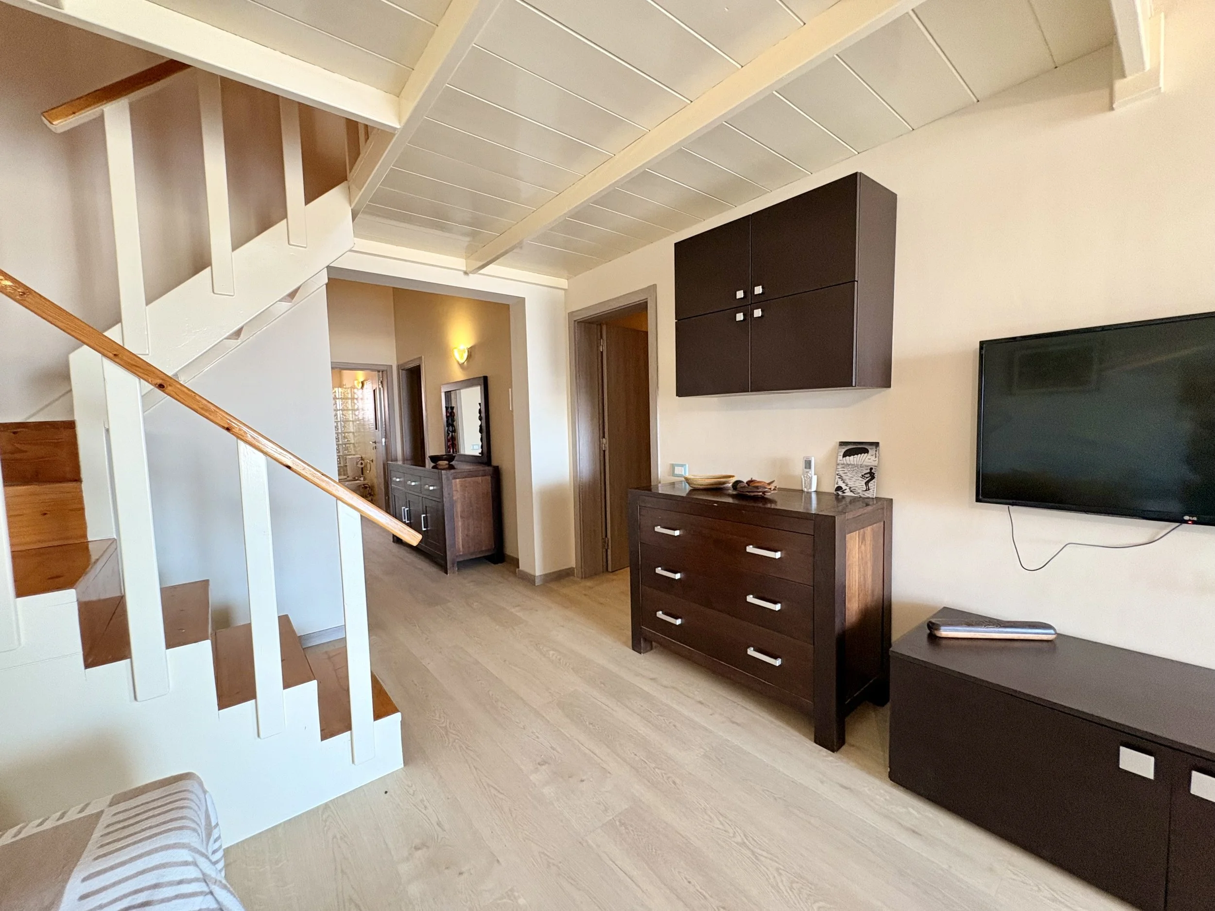 Living room area with a wooden dresser, a wall-mounted cabinet, a flat-screen TV, and a staircase with wooden steps and white railing.