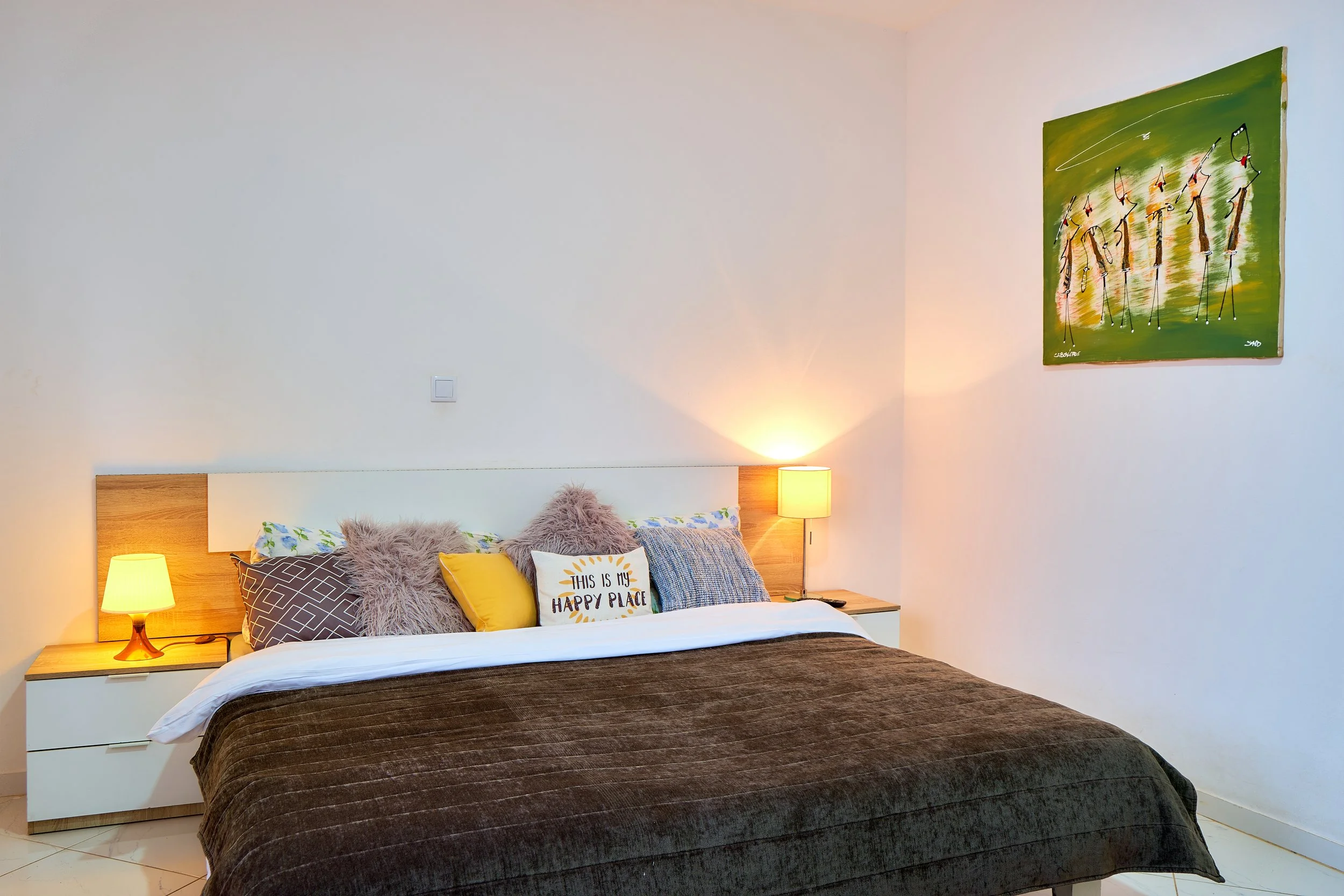 A bedroom with a bed featuring a dark brown blanket, multiple pillows including a yellow one and a pillow that says 'This is my happy place,' wooden nightstand with a yellow lamp, white wall with an abstract painting, and a light switch.