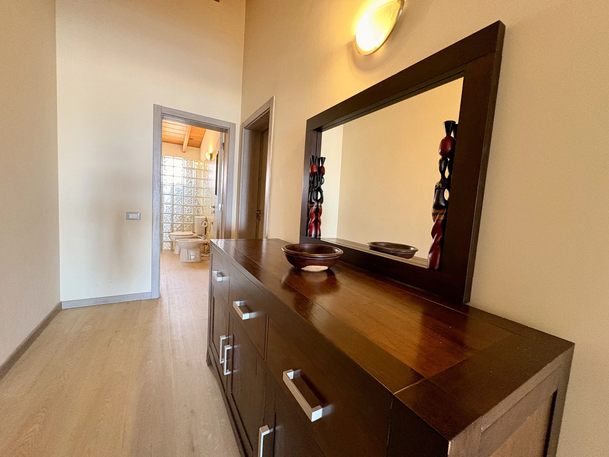 A hallway with a hardwood dresser topped with decorative bowls and a mirror, leading to a bathroom with glass block windows and a toilet.