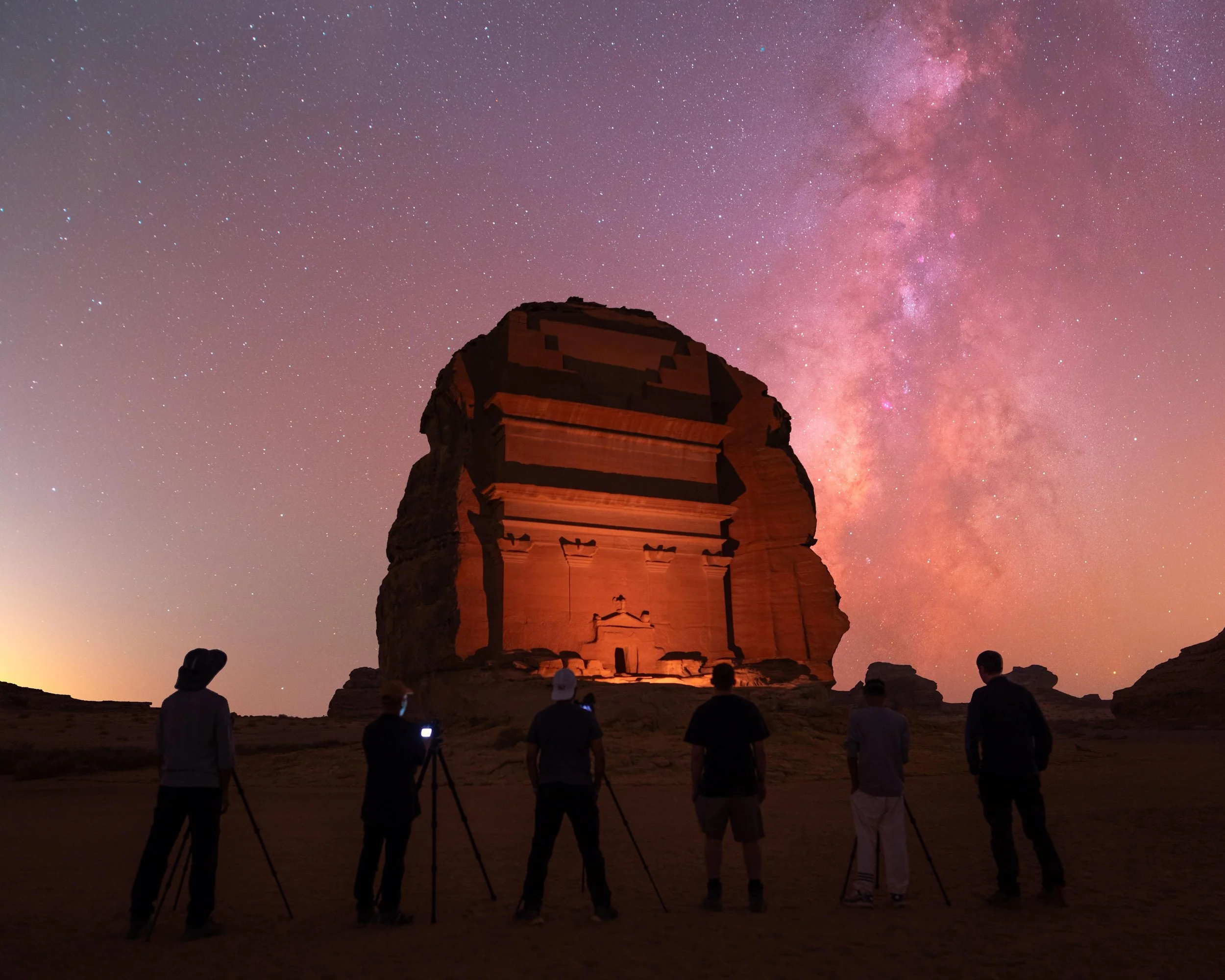 People are observing the night sky and taking photographs of the Milky Way above an ancient archaeological site with stone structures.