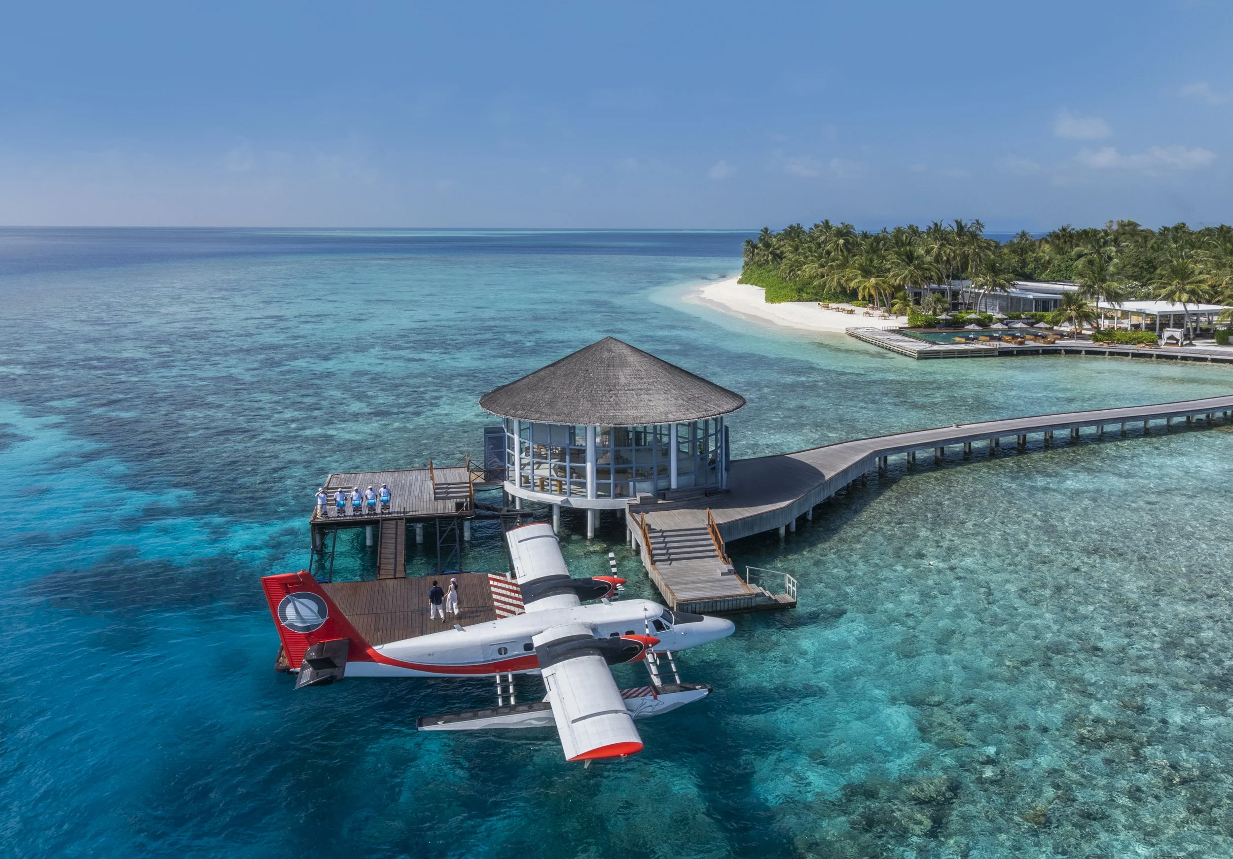 Aerial view of a seaplane docked at a tropical island resort with clear blue waters, a white sandy beach, and lush green palm trees.