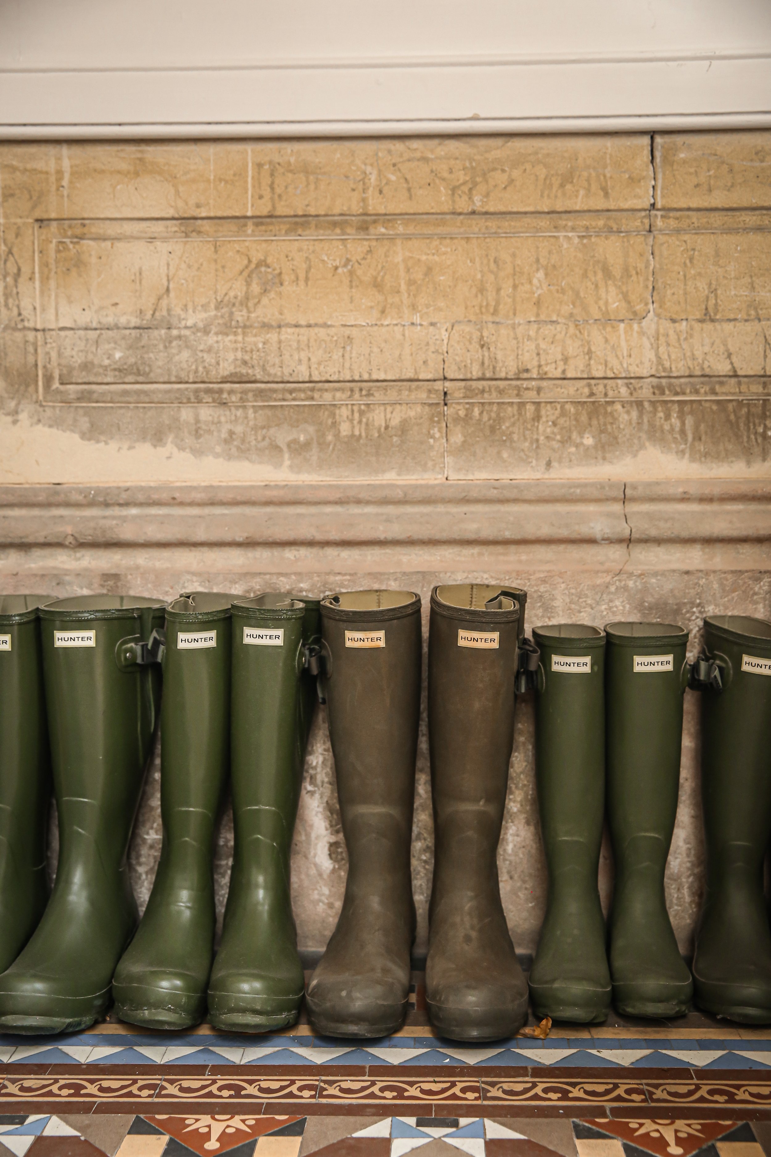 Group of green rubber rain boots with labels reading 'HUNTER' lined up against a stone wall with patterned tile floor in the background.