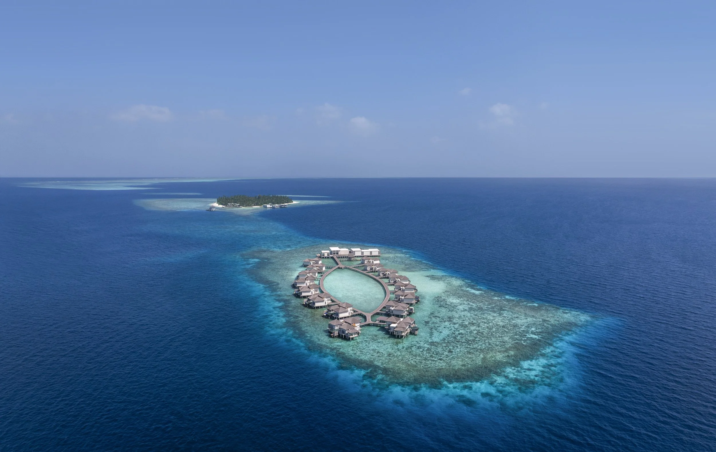 Aerial view of an overwater bungalow resort on a tropical island with turquoise water and clear skies.