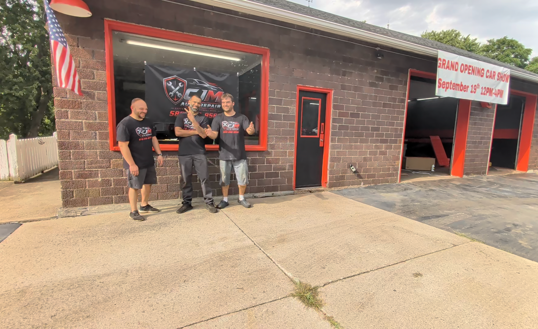 Three men stand outside a brick building with a large window, a door, and a banner advertising a grand opening event. They are smiling and appear to be celebrating.
