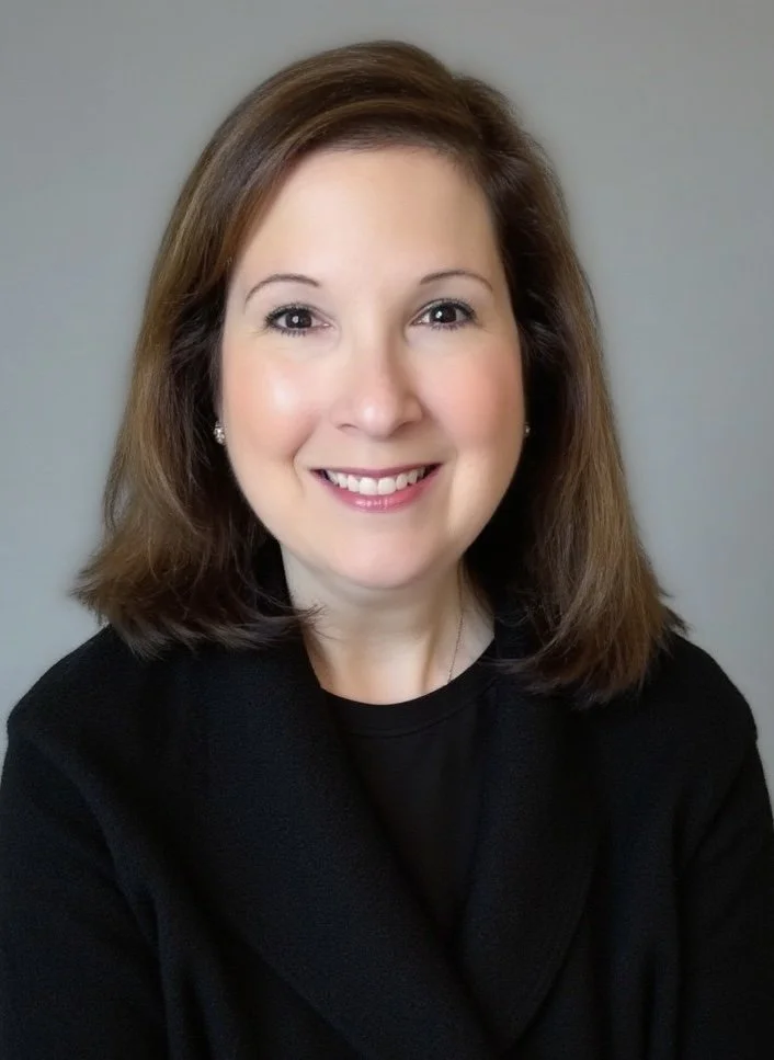 A woman with shoulder-length brown hair smiling at the camera, wearing a black top and earrings, against a plain gray background.