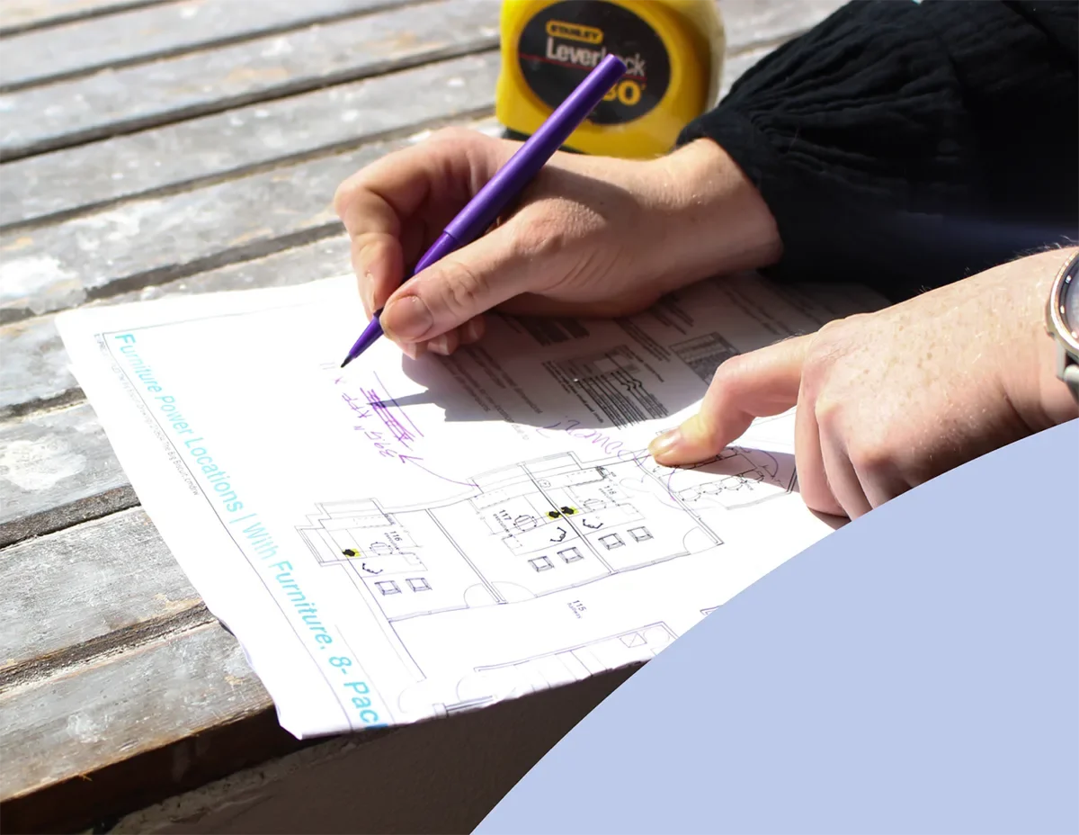 Hands marking a floor plan for furniture placement with a purple pen beside a tape measure