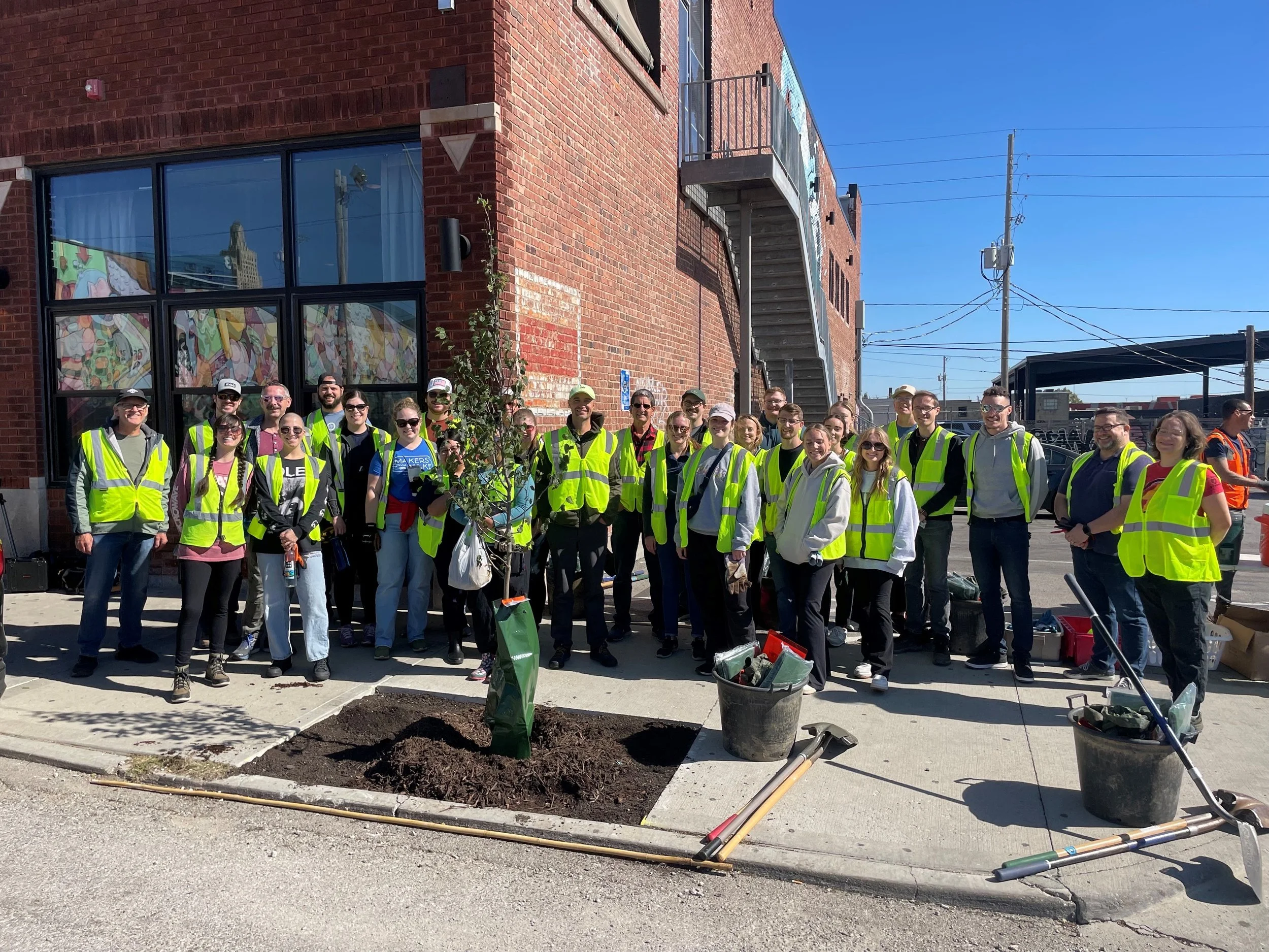 Community volunteers in safety vests pose beside a newly planted street tree, gardening tools, and soil outside a brick building.