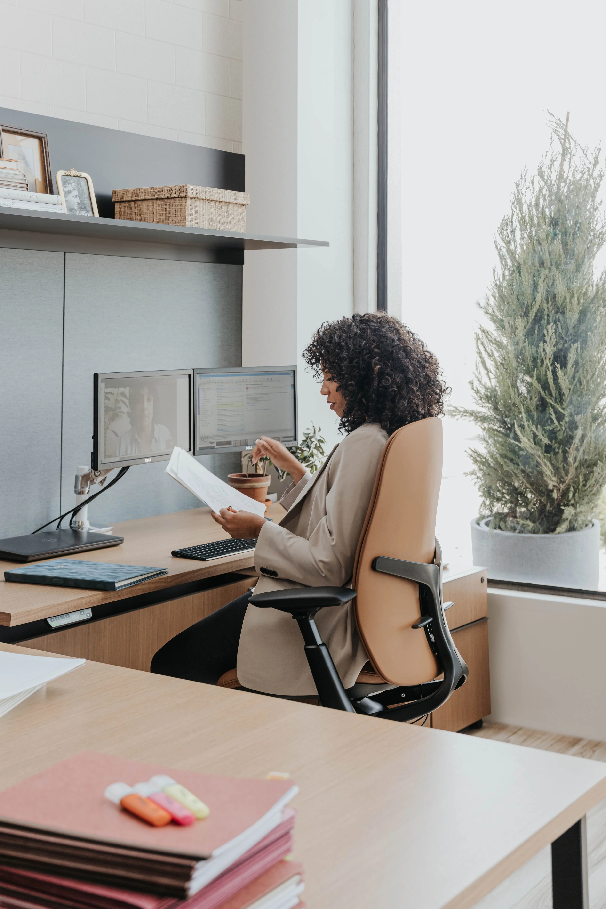 Professional woman reviewing papers at a dual-monitor home office desk beside a large window.