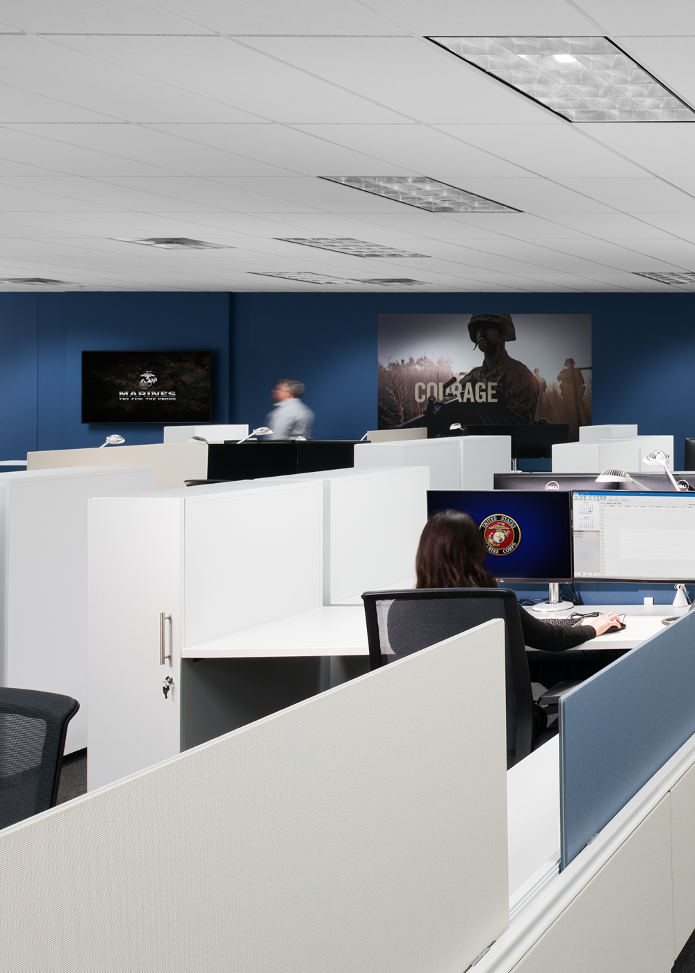 Modern open-plan office with cubicles, employees at computers, and U.S. Marine Corps posters on blue walls