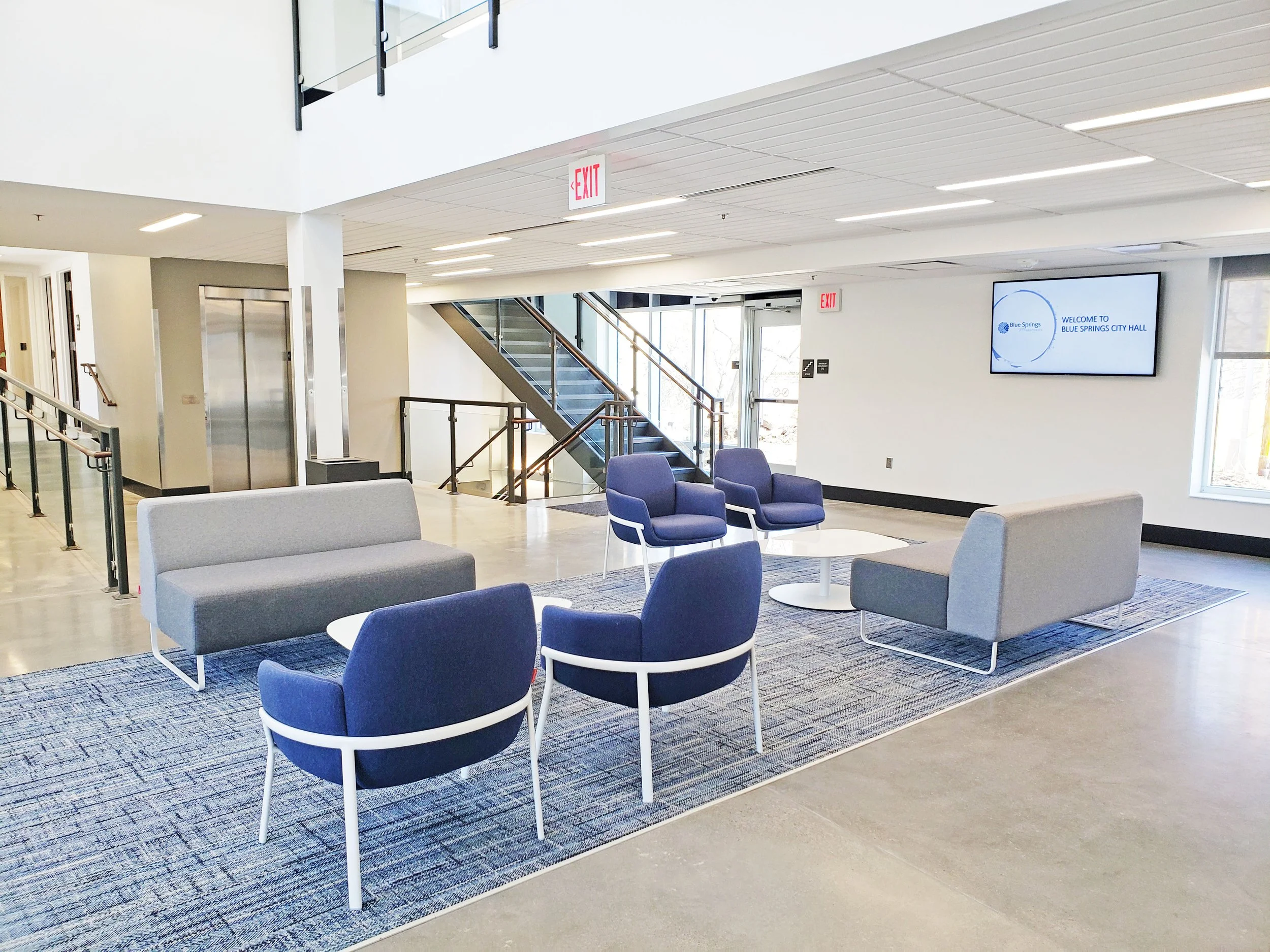 Modern Blue Springs City Hall lobby with blue and gray seating, staircase, elevator, exits, and welcome display