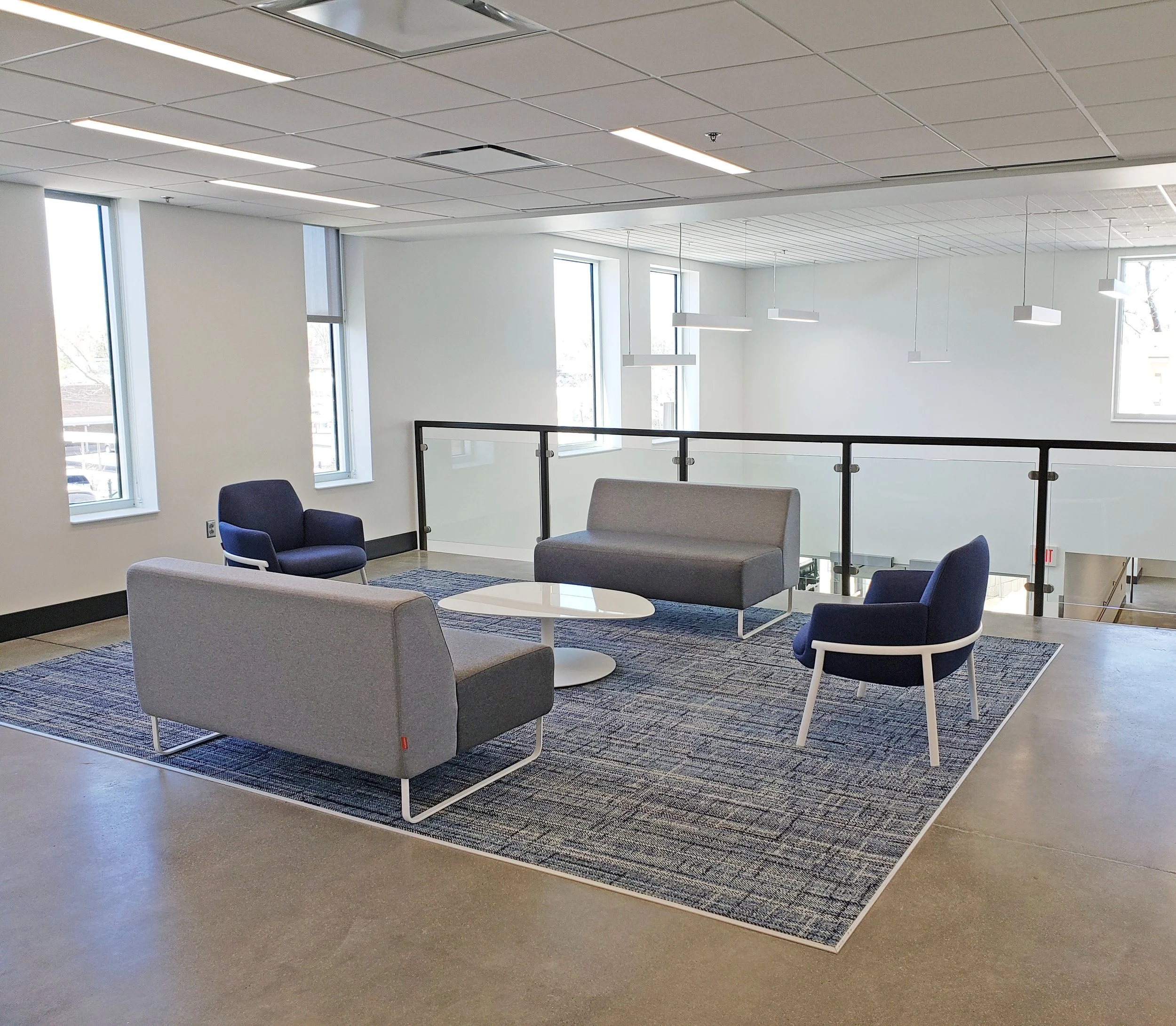 Modern office lounge with gray sofas, blue armchairs, white coffee tables, patterned rug, pendant lights, and glass railing.