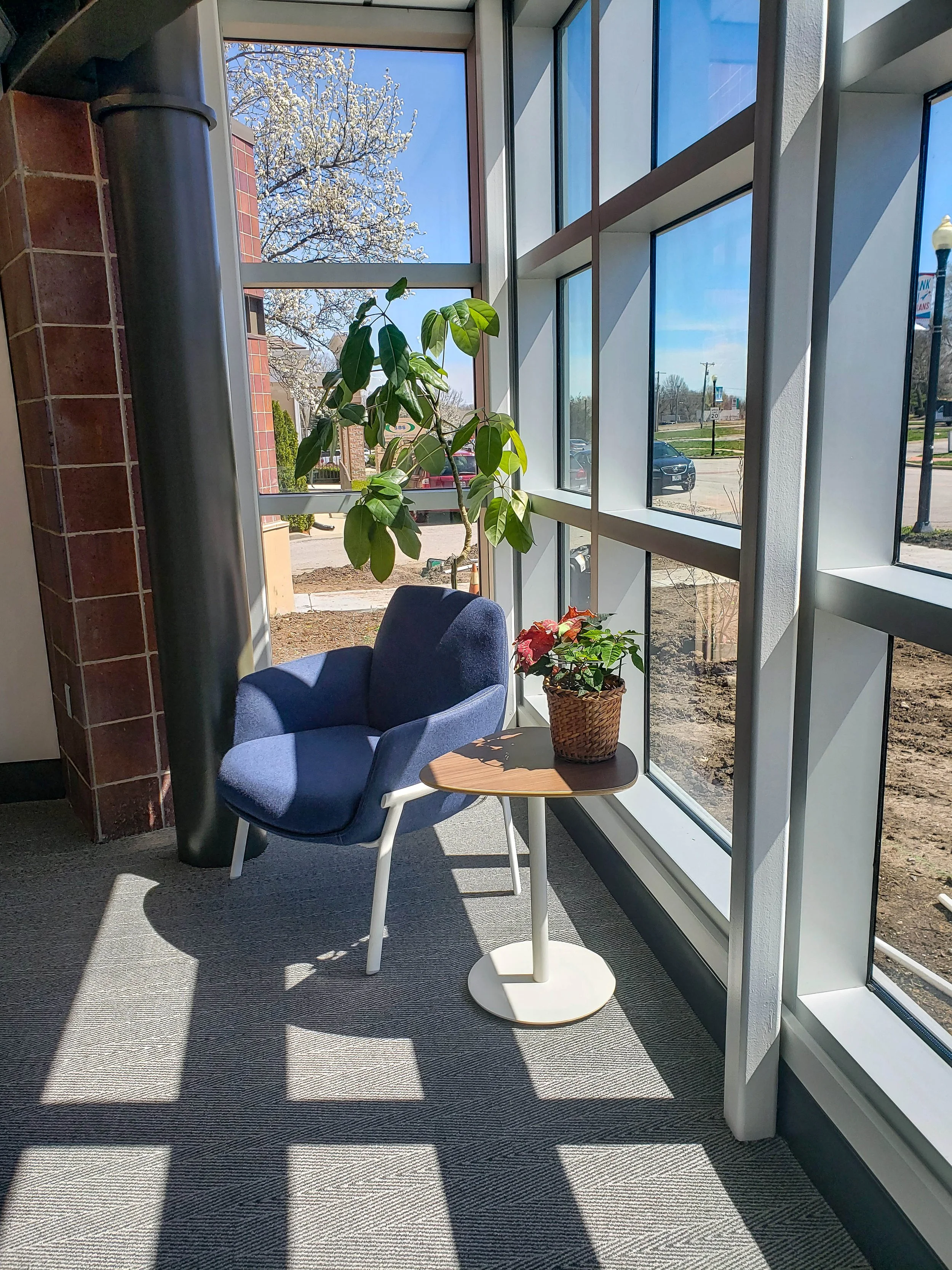 Bright office sitting area with a blue armchair, round table, potted plants, and floor-to-ceiling windows overlooking a street.