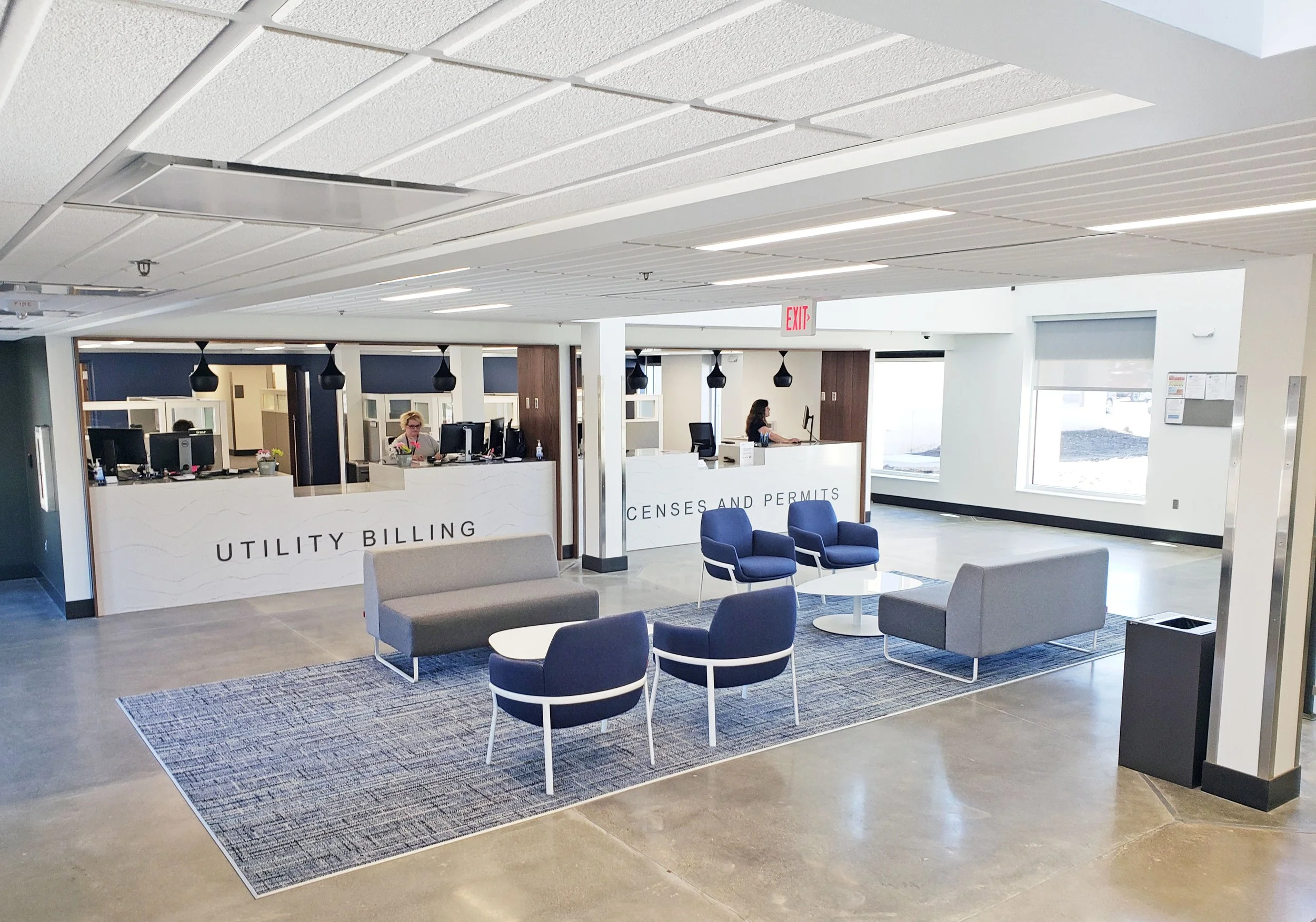 Modern municipal office lobby with utility billing and census and permits service counters, waiting chairs, and an exit sign.