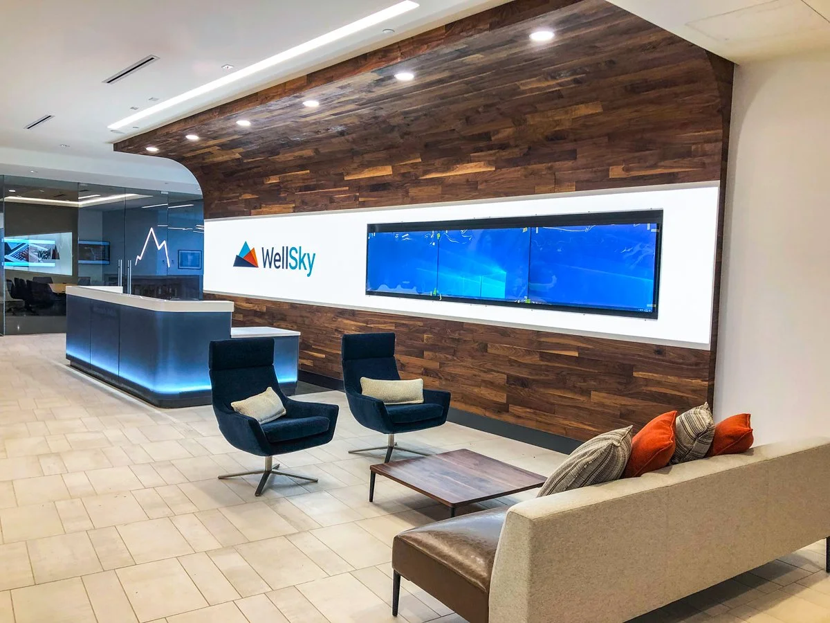 Modern WellSky office reception area with wood-paneled wall, illuminated logo, digital display, reception desk, and lounge seating