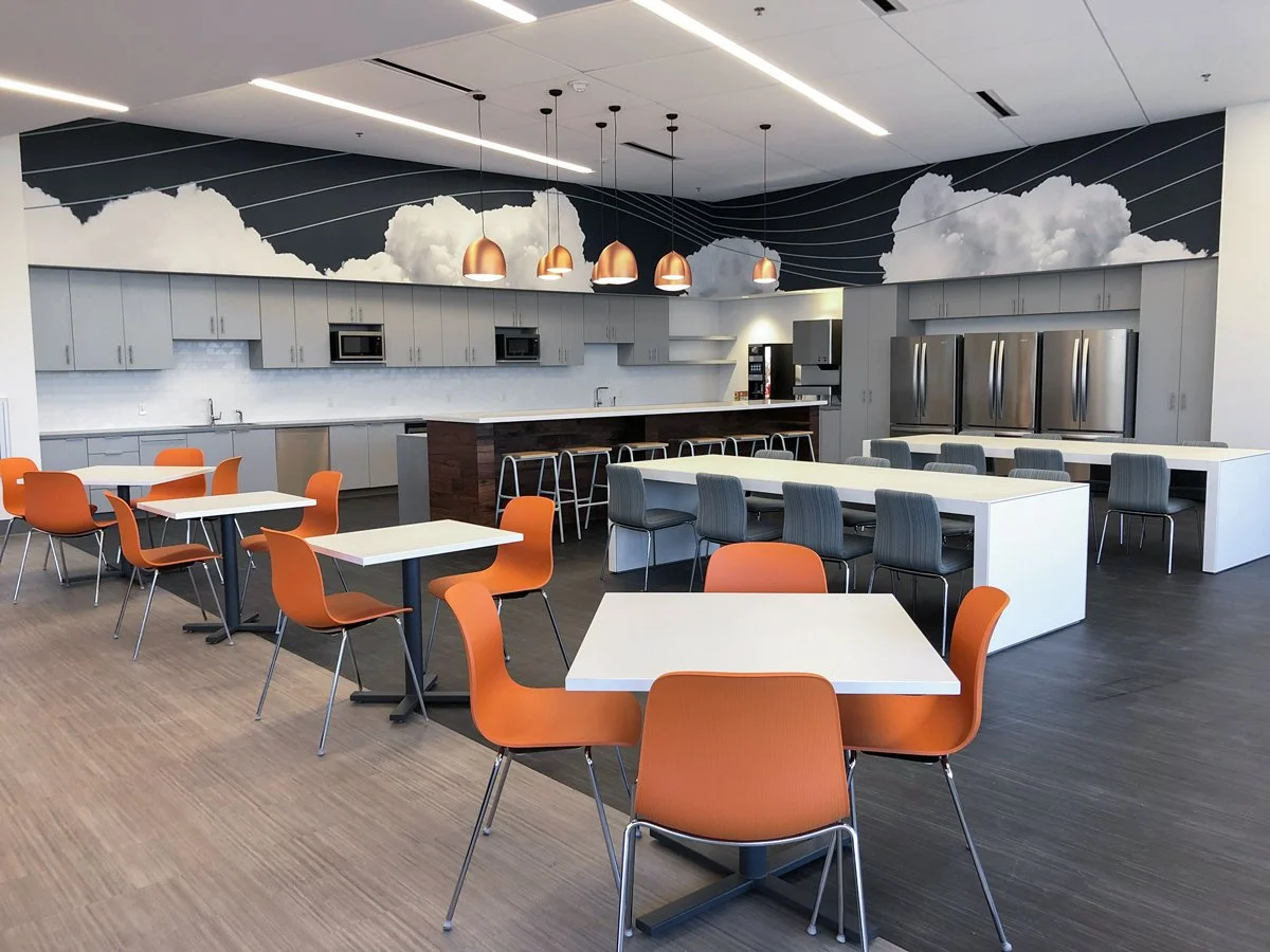 Modern office break room with orange and gray chairs, dining tables, kitchenette, stainless steel refrigerators, and cloud mural