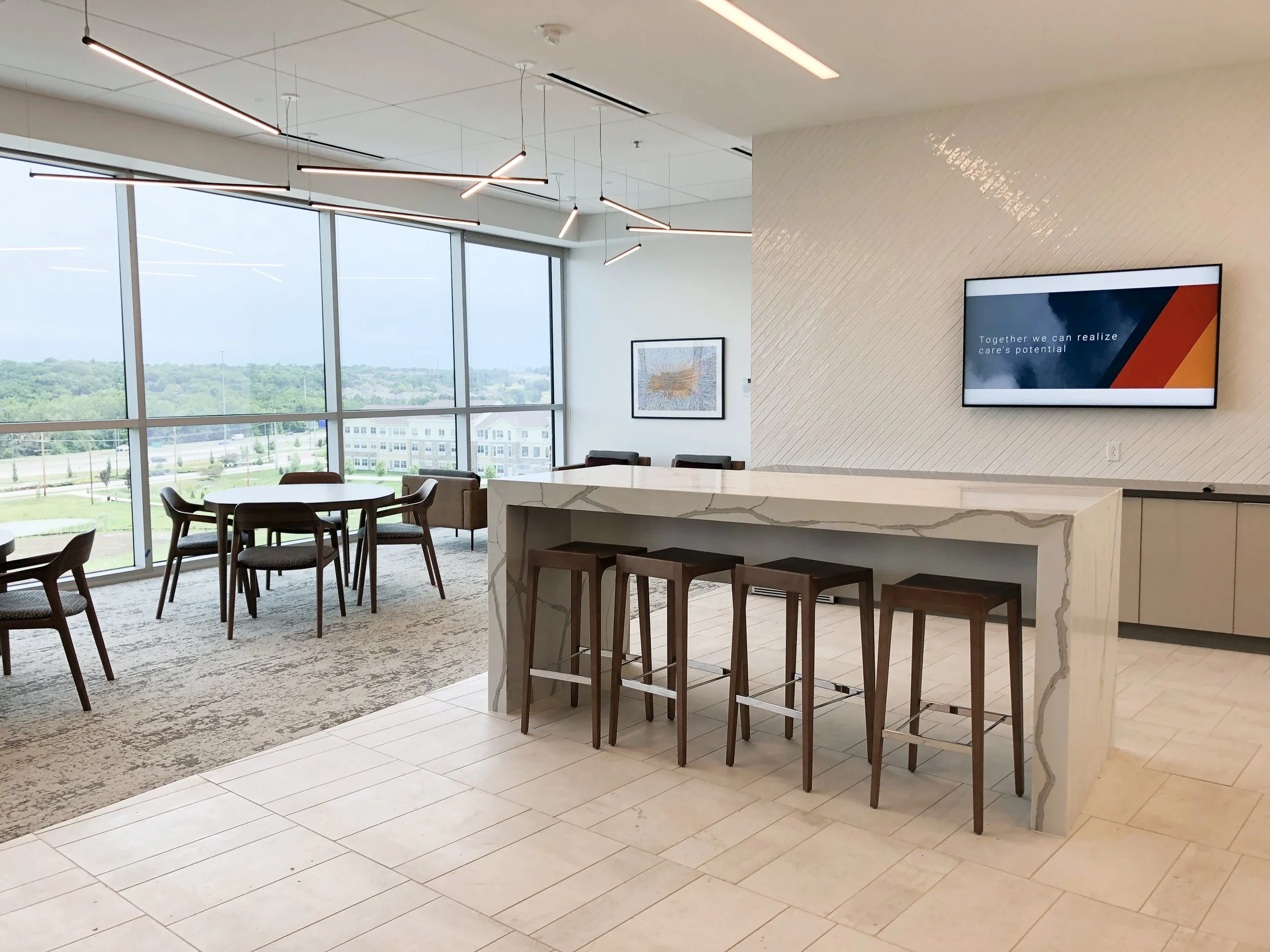 Modern office lounge with a marble island, bar stools, dining tables, pendant lights, large windows, and a wall-mounted display