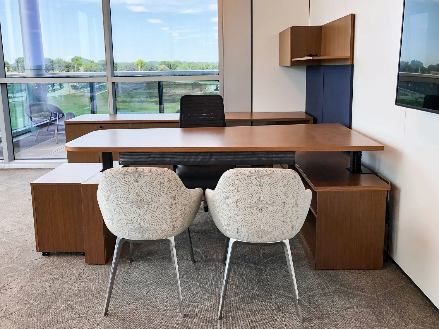 Modern office workstation with a wooden desk, two patterned guest chairs, an office chair, storage cabinets, and large windows