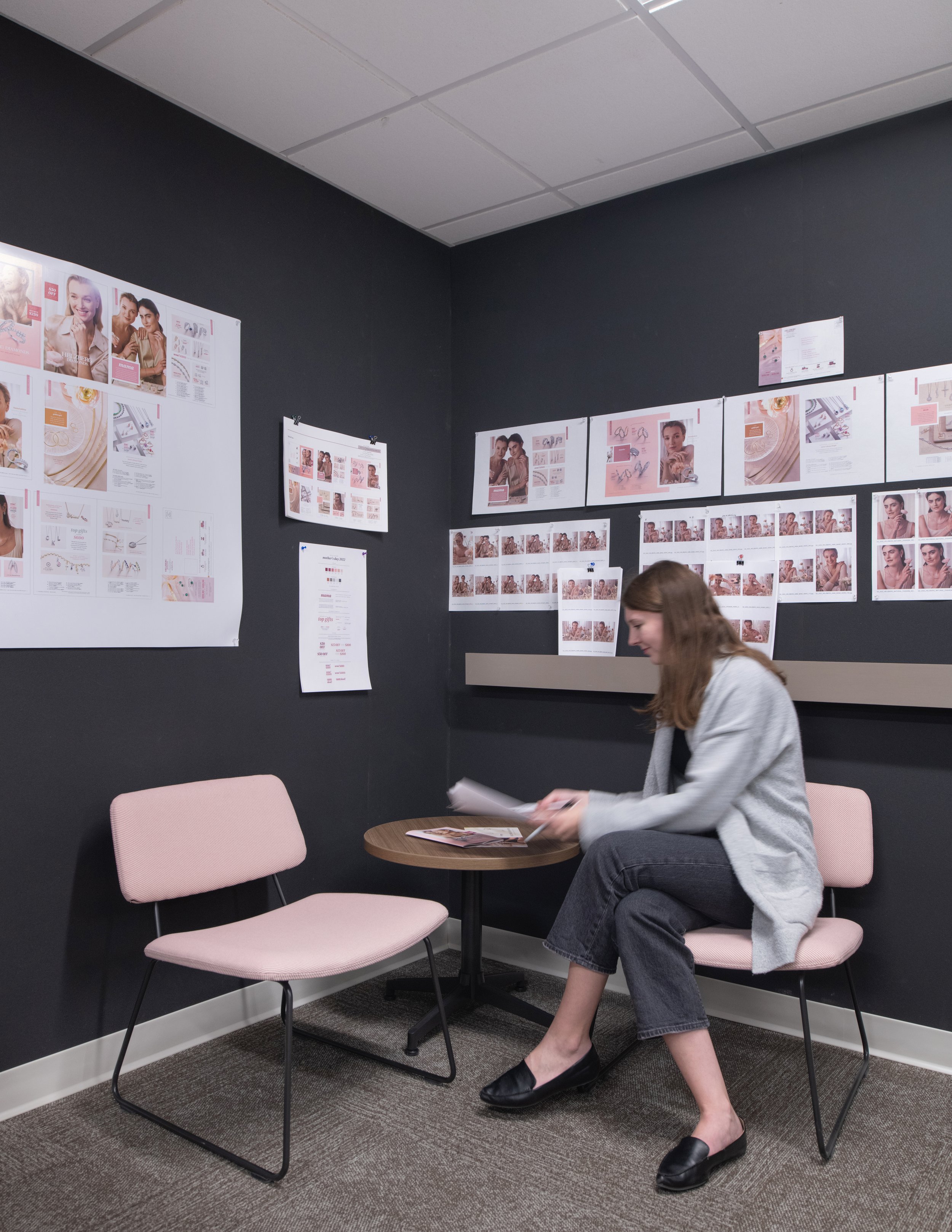 Woman reviewing jewelry design boards in a studio meeting room with pink chairs and a round table