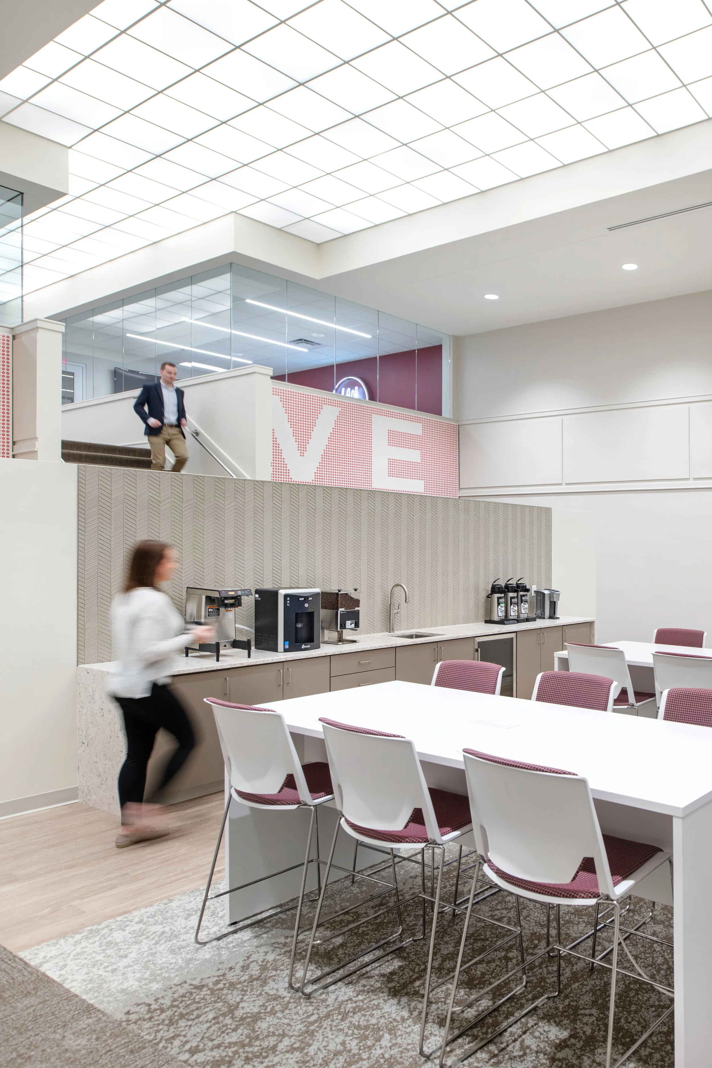 Modern office break room with coffee and beverage stations, dining tables, white chairs, glass walls, and a person walking downstairs