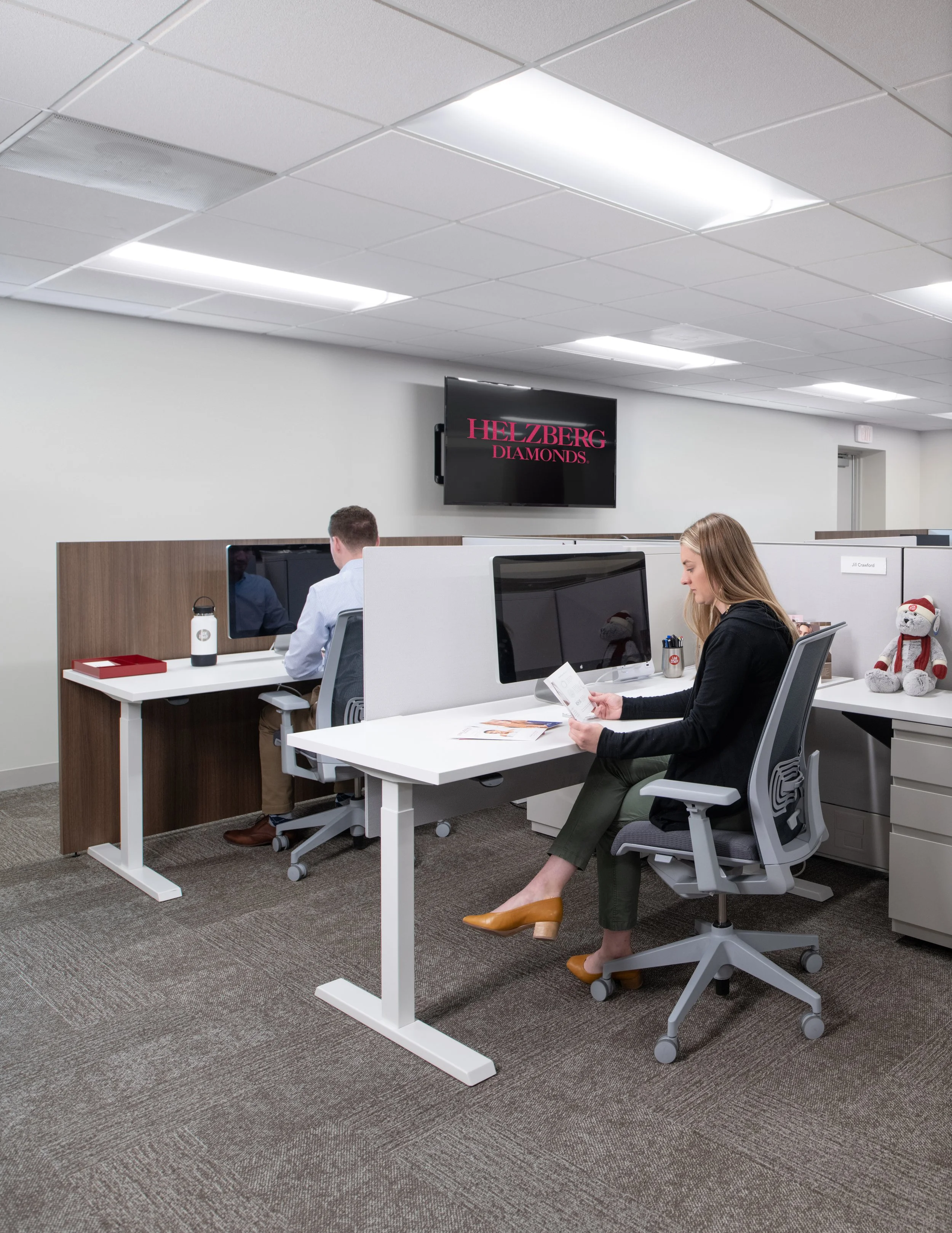 Employees working at desks in a modern office with computer monitors and a Helzberg Diamonds sign