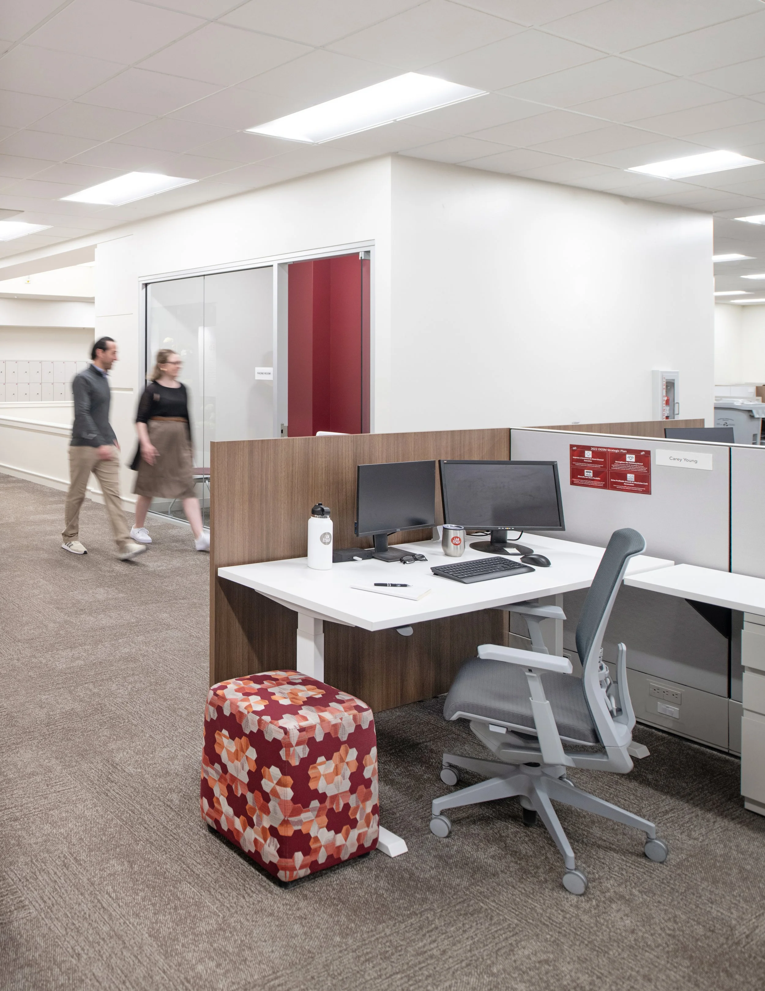Modern open-plan office with computer workstations, desks, office chairs, lockers, and two coworkers walking past a phone room.