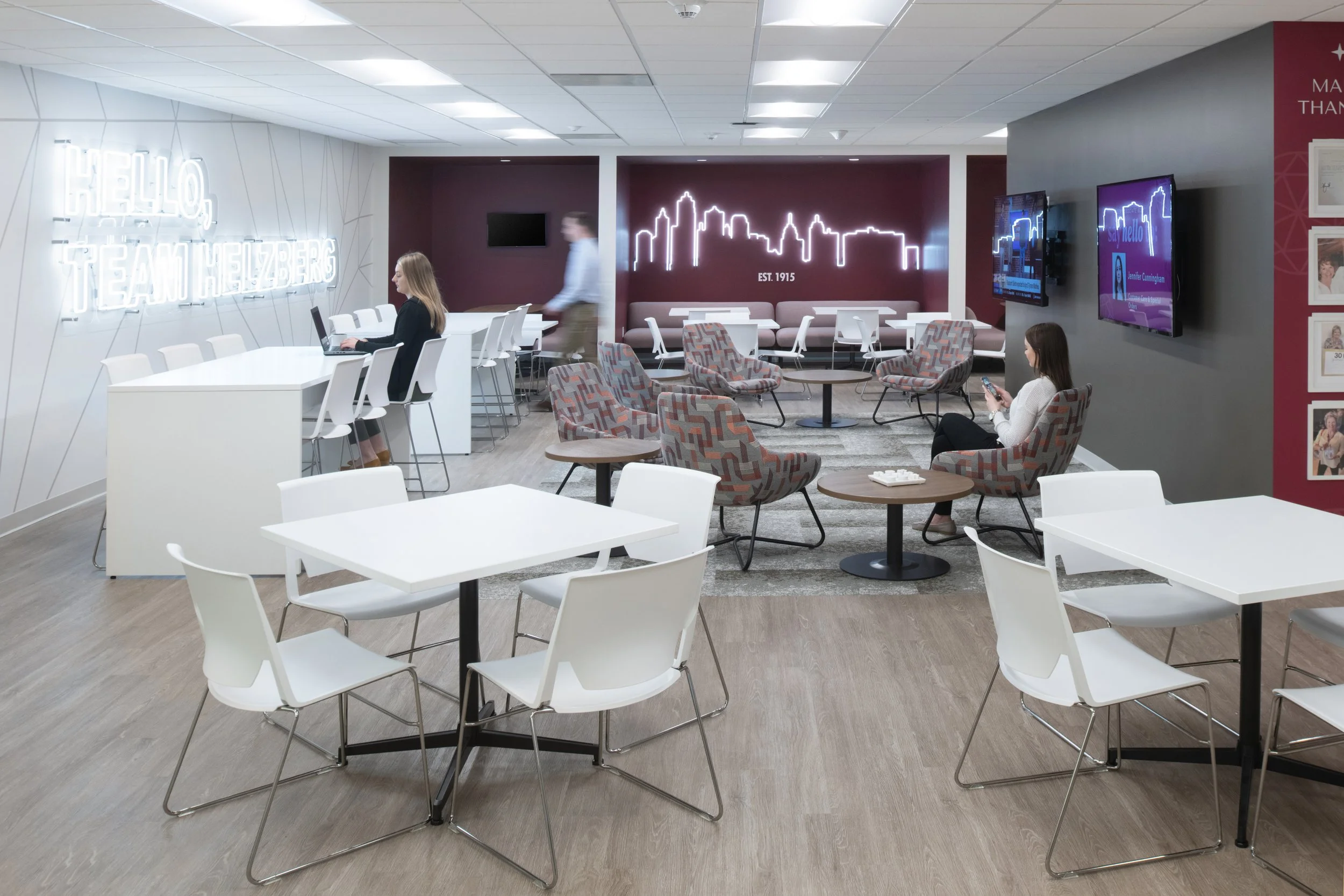 Modern corporate lounge with white tables, patterned chairs, neon skyline artwork, televisions, and employees working or relaxing