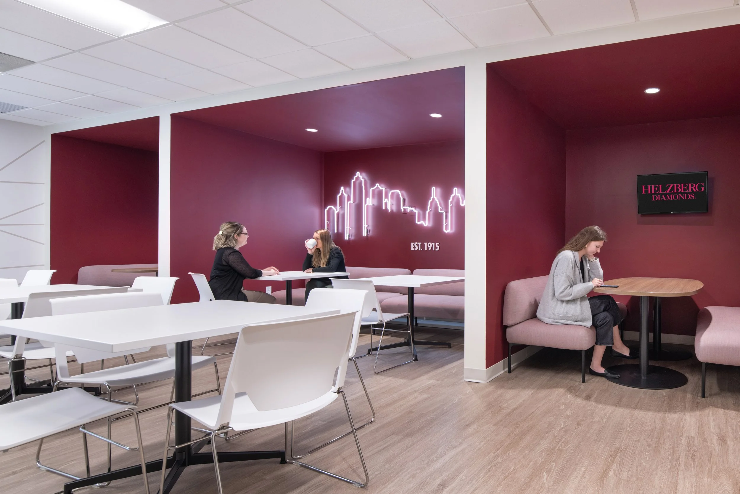 Modern Helzberg Diamonds lounge with burgundy walls, white tables and chairs, pink seating, neon city skyline sign, and three women.