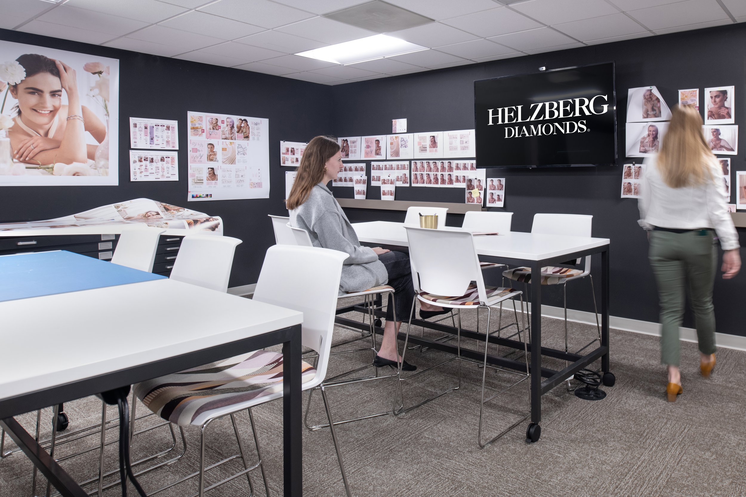 Helzberg Diamonds design studio with two women, jewelry campaign mood boards, display tables, and a branded wall monitor