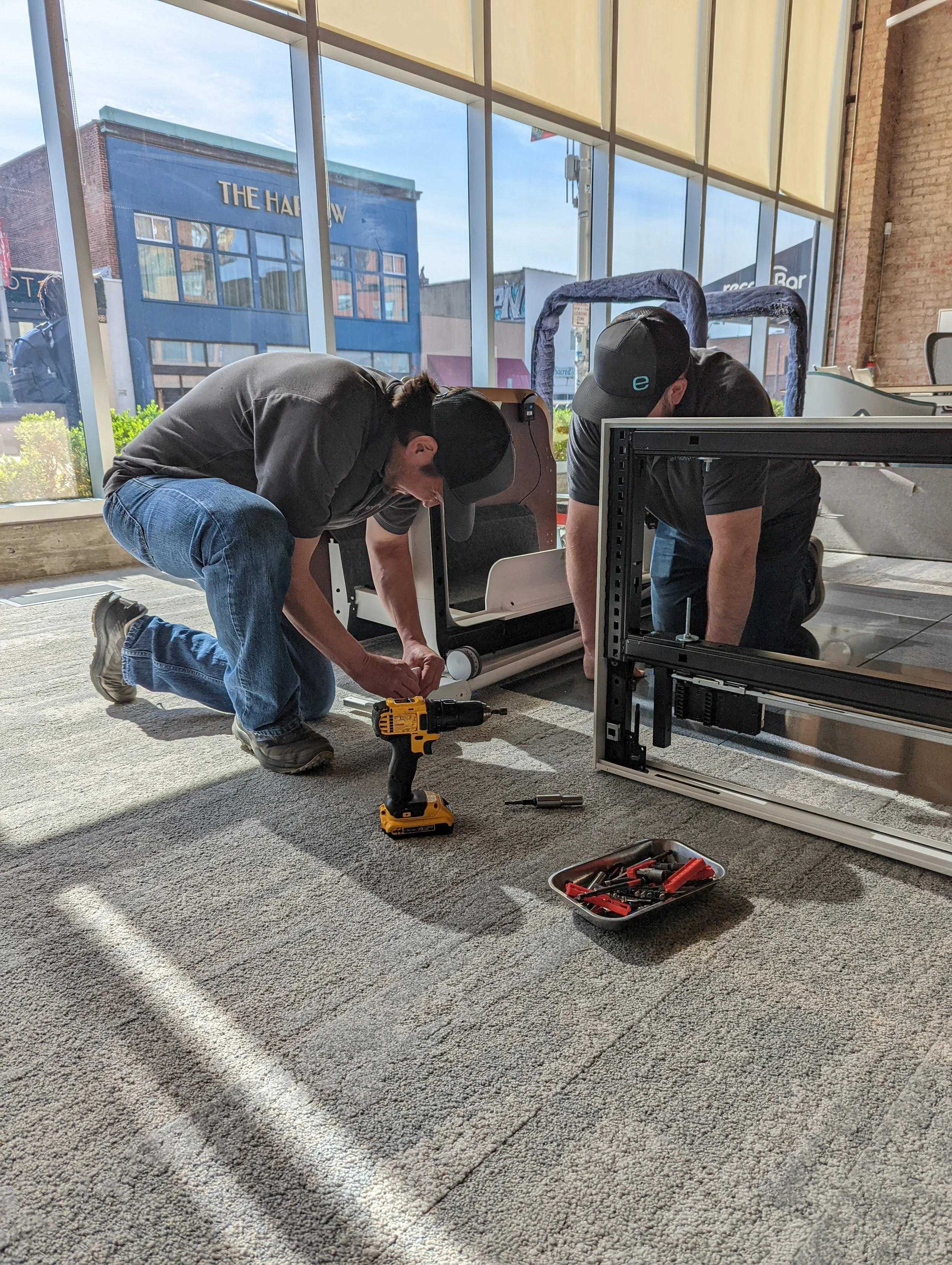 Two technicians in a storefront install or repair equipment beside a computer rack, with power tools and a tool tray on the carpet.