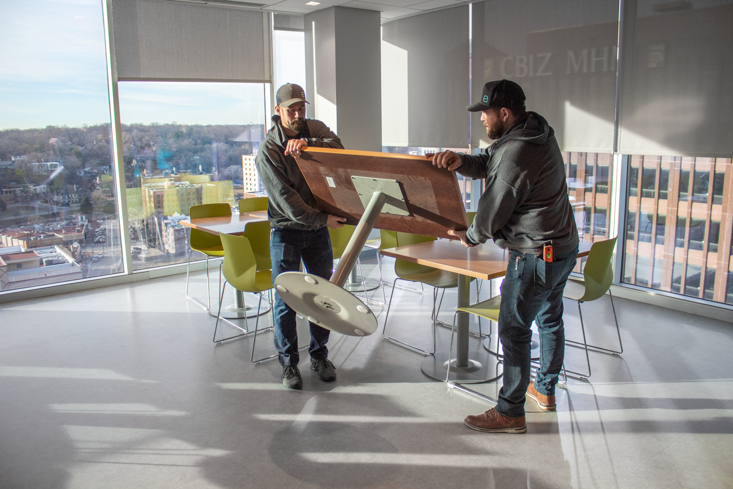 Two men move a large tabletop in a sunlit office lounge with floor-to-ceiling windows and city views.
