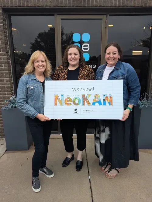 Three women holding a welcome sign reading “Neokani” outside a building