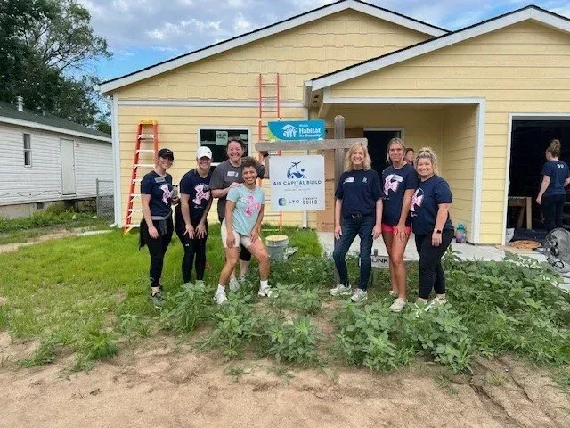 Habitat for Humanity volunteers pose outside a yellow house under construction.