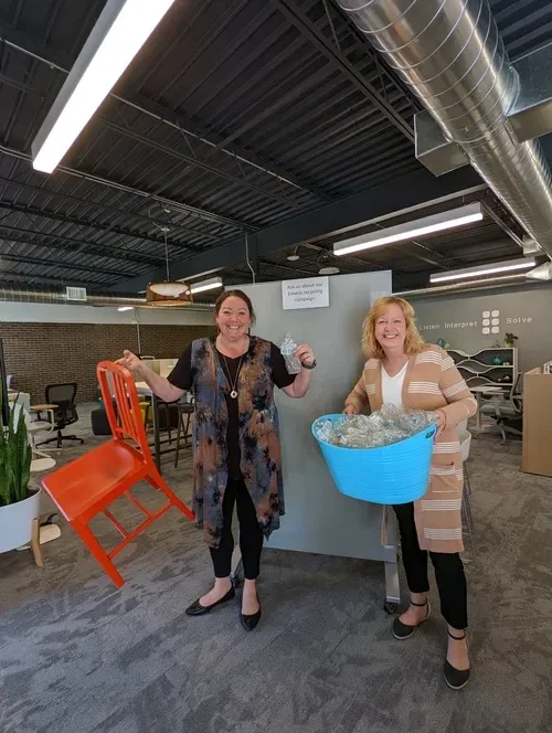 Two smiling women in an office holding a red folding chair and a blue basket filled with glass items.