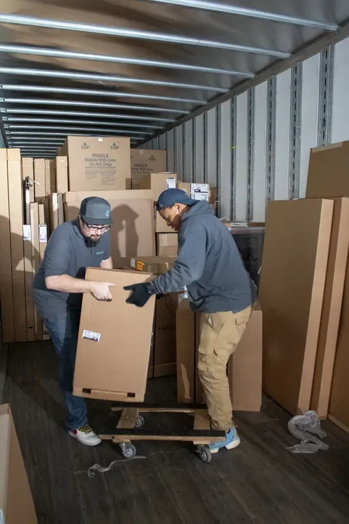 Two warehouse workers moving a large cardboard box on a dolly inside a loaded shipping trailer