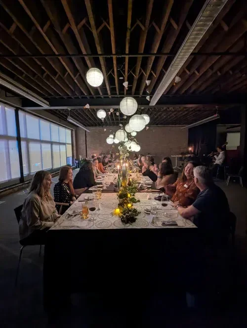 Long dinner table with guests dining beneath hanging globe lights in a rustic industrial event space