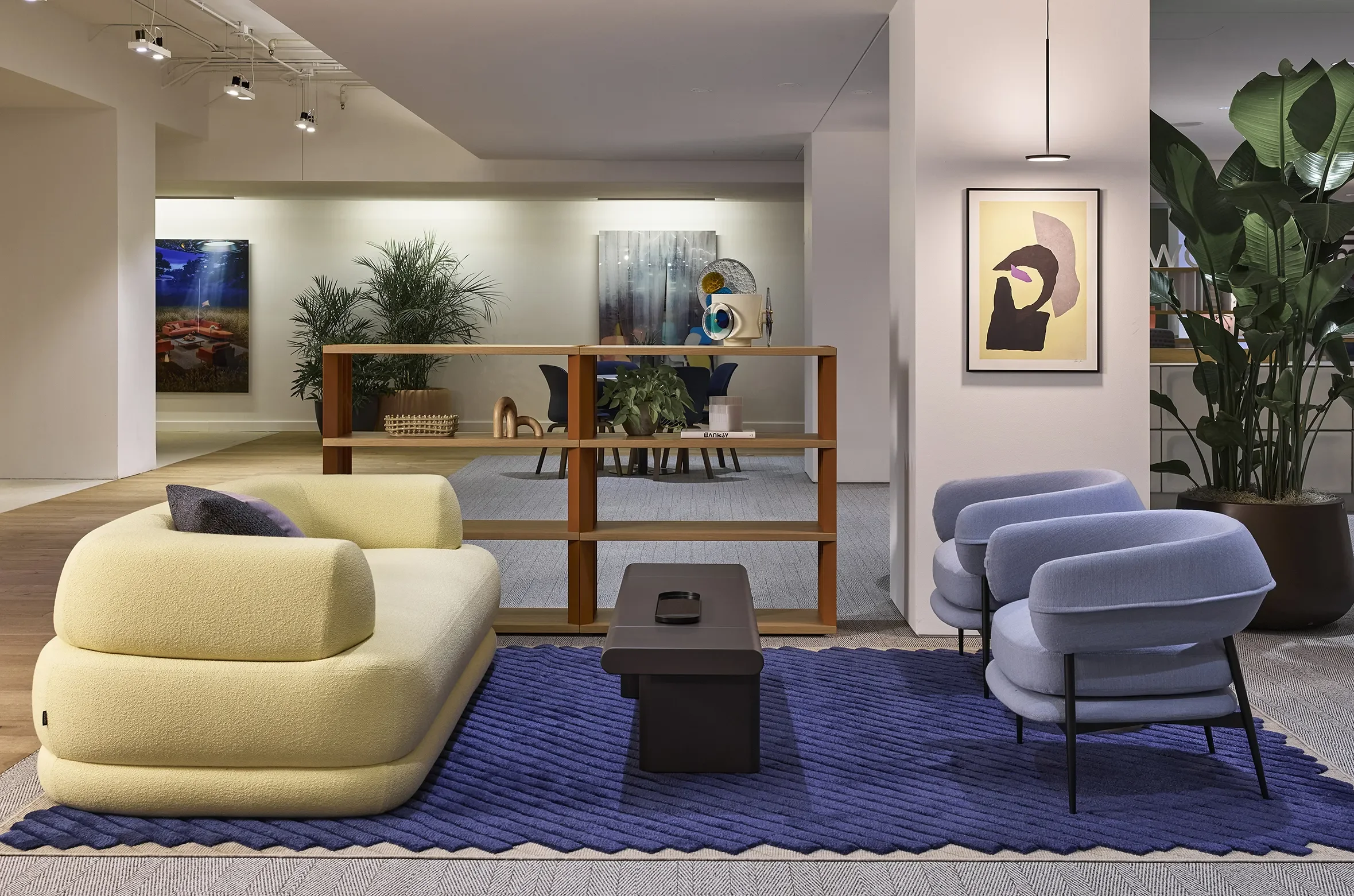 Modern office lounge with yellow and lavender chairs, a wooden shelving divider, plants, artwork, and a blue textured rug