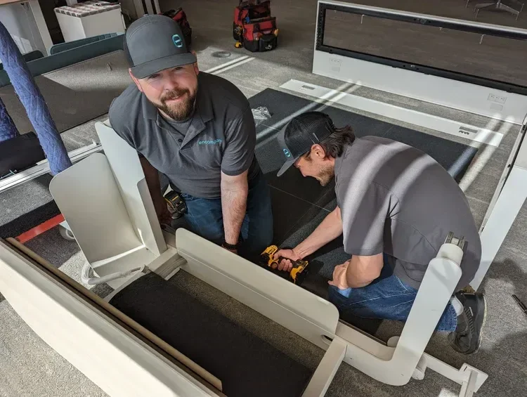 Two technicians wearing gray uniforms repair a commercial treadmill with a power drill.