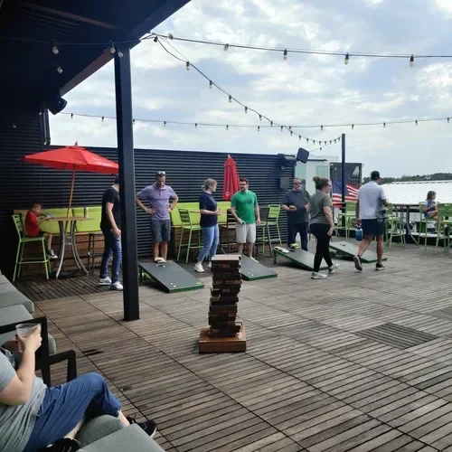 People playing cornhole on a waterfront deck with string lights, red umbrellas, and a Jenga tower