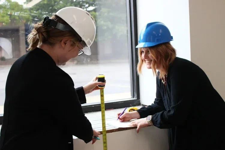 Two women in hard hats measuring and marking a windowsill during a construction inspection