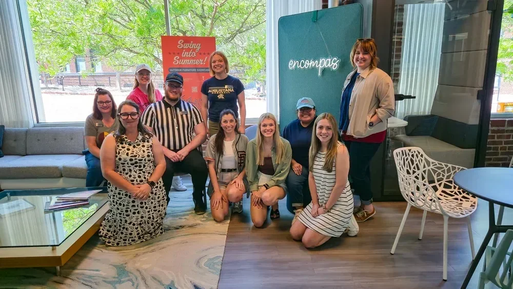 Group of 11 people posing inside a lounge beside an Encompass sign and Summer concert poster
