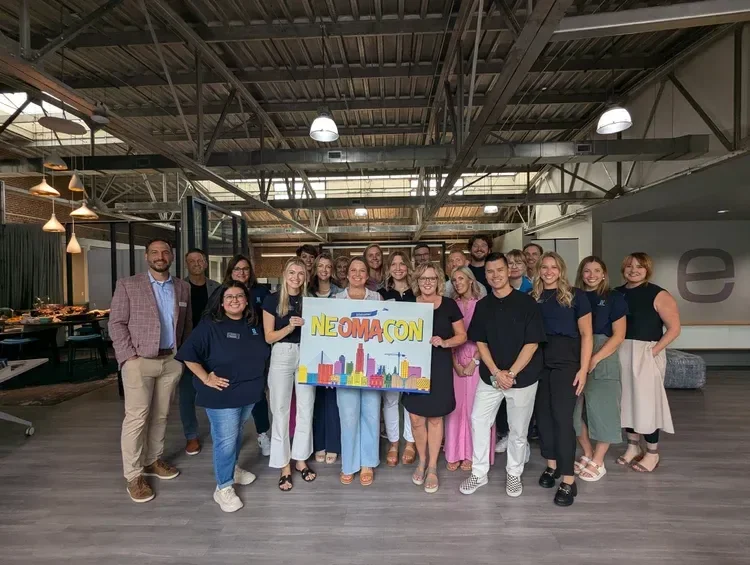 Group of coworkers holding a colorful NEOMACON sign in a modern office